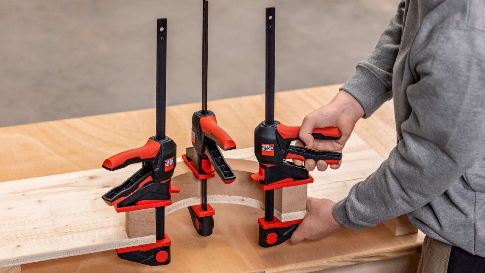 A person uses three red and black clamps to secure wooden boards together on a workbench, demonstrating a woodworking or carpentry process.