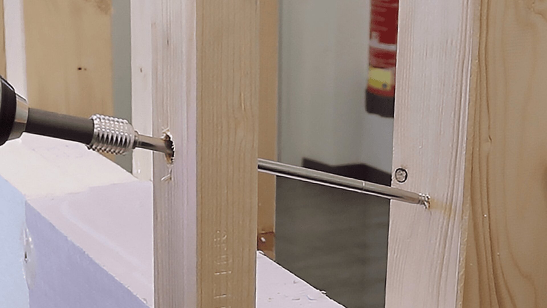 A drill is driving a long screw through two wooden studs, fastening them together. A fire extinguisher is visible in the blurry background.