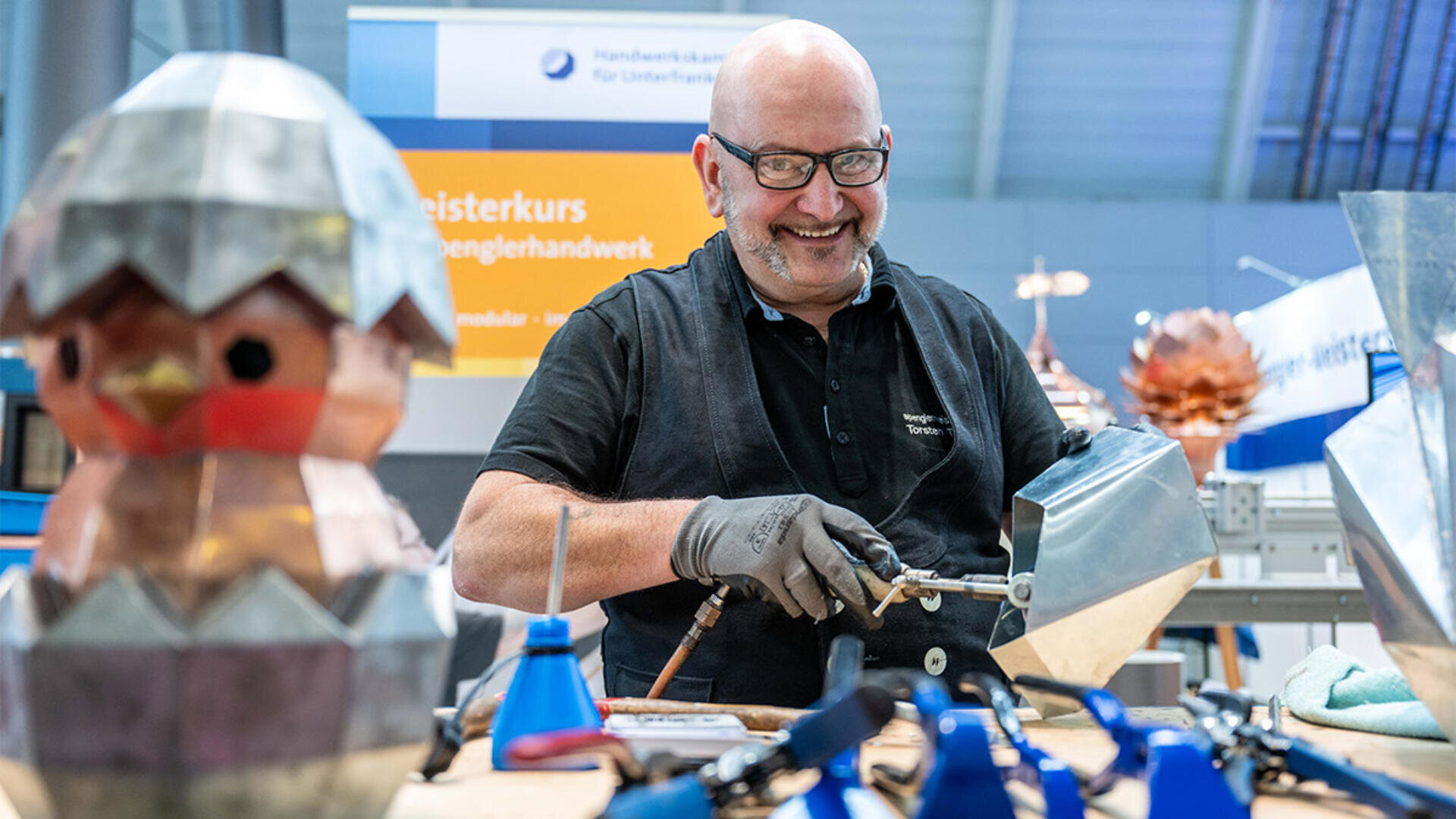 Ein lächelnder Mann mit Brille und Handschuhen verwendet ein Schweißgerät an einem Metallobjekt an einer Werkbank, umgeben von Metallskulpturen und Werkzeugen in einer Werkstattumgebung.