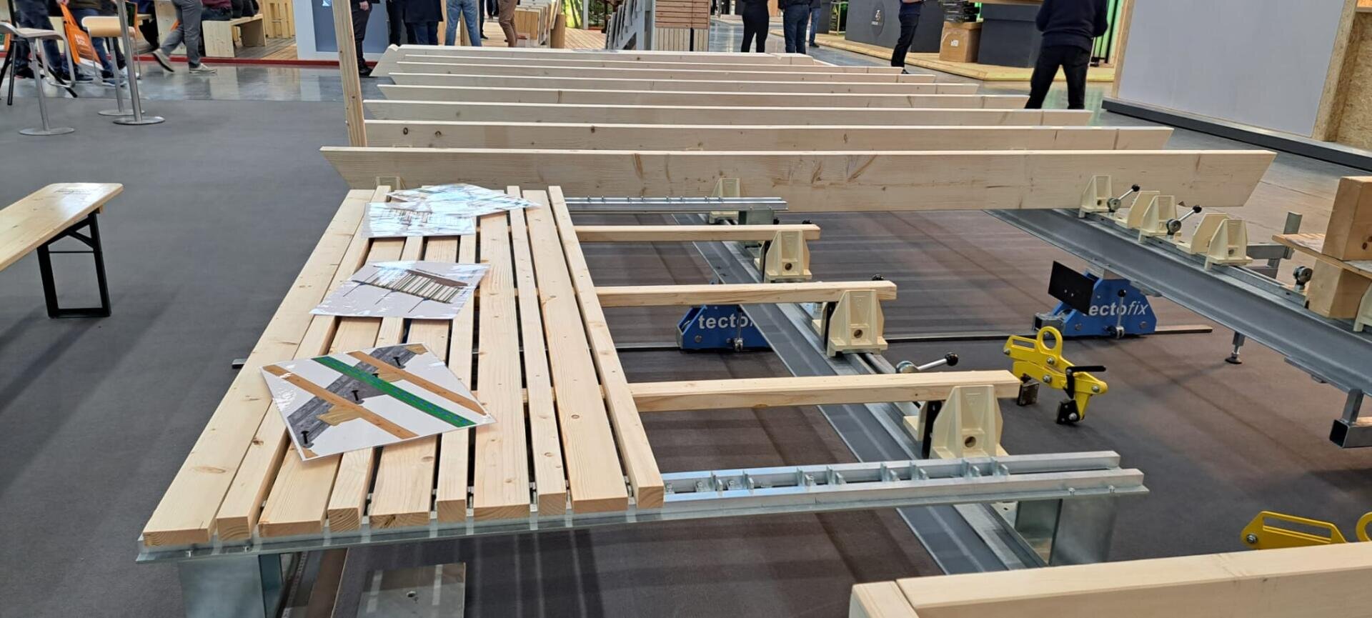 Mehrere Holzbretter und -balken sind auf einem Metallrahmen in einem Innenraum angeordnet, darauf befinden sich Baupläne und Flugblätter. Im Hintergrund sind Menschen zu sehen. Die Szene scheint Teil einer Bau- oder Handelsausstellung zu sein.