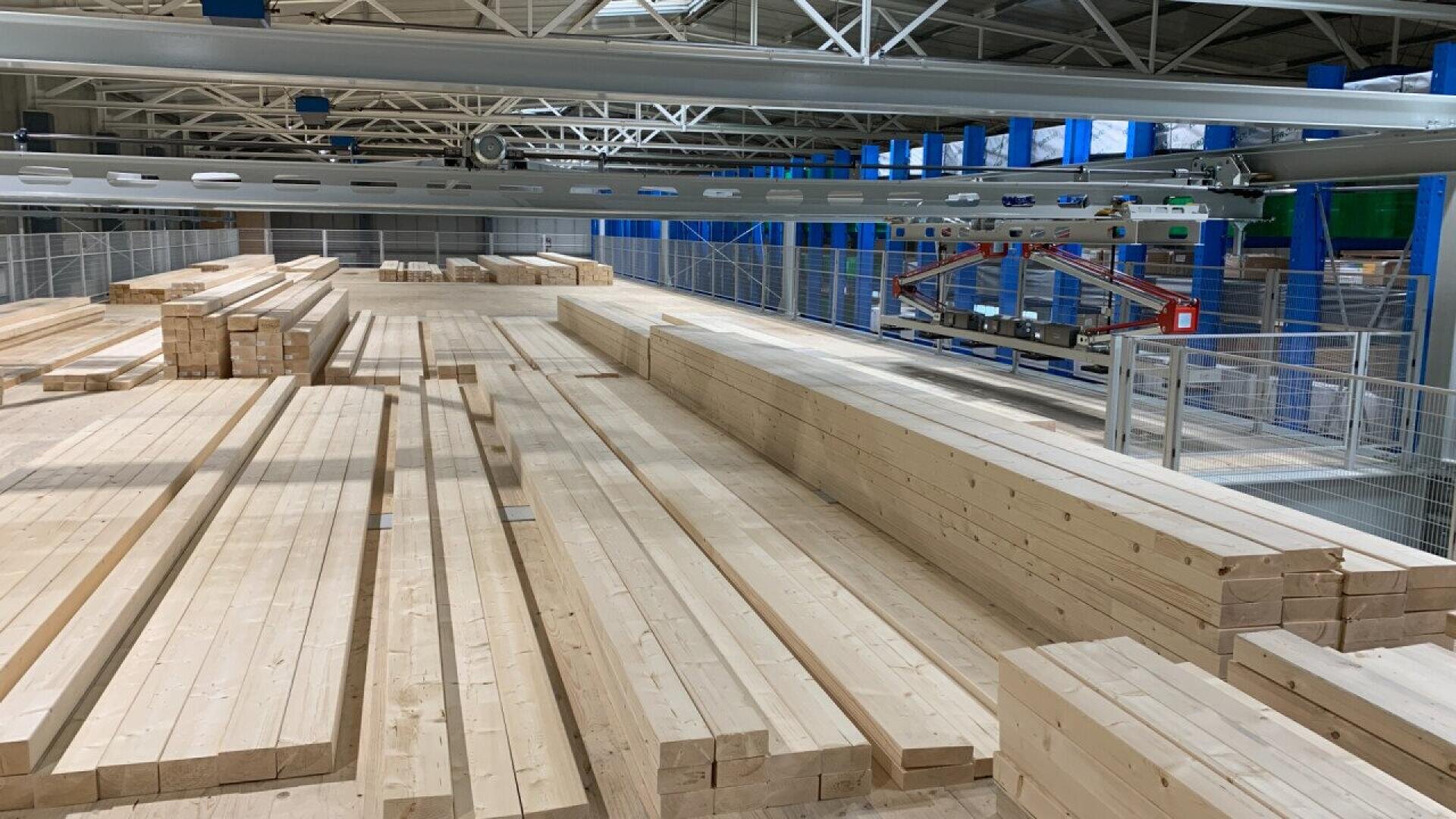 Stacks of neatly arranged wooden beams are lined up in a spacious, well-lit warehouse with metal railings and a robotic machine overhead for handling the timber.