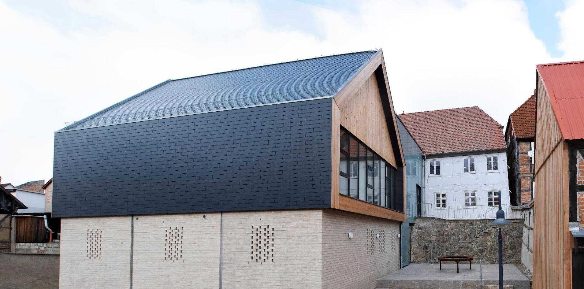 Modernes Gebäude mit einem beigen Ziegeluntergeschoss und einer schwarzen, schrägen Oberfassade. Große Fenster säumen die Seite, und es ist von älteren Häusern mit roten Dächern umgeben. Im Vordergrund befinden sich eine Steinmauer und ein kleiner Innenhof.