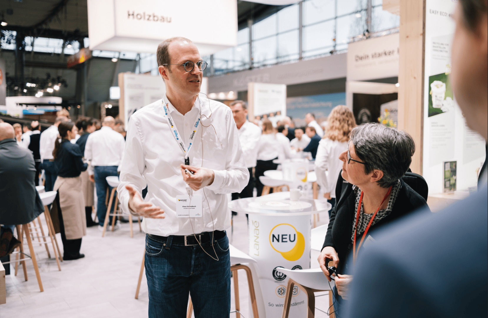 Ein Mann mit weißem Hemd und Brille spricht mit einer Frau in einer belebten Ausstellungshalle, in der im Hintergrund andere Menschen und Informationsstände zu sehen sind. Die Atmosphäre wirkt professionell und lebendig.