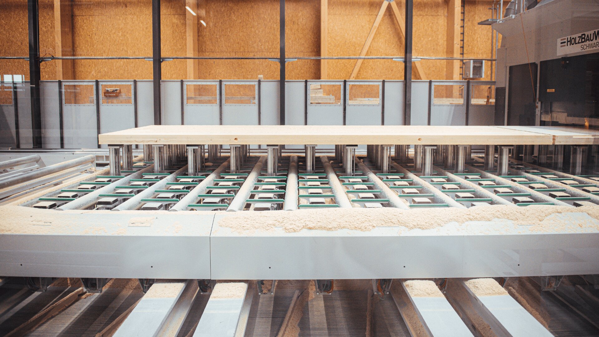 Eine große industrielle Holzbearbeitungsmaschine in einer Fabrik bearbeitet eine dicke Holzplatte, auf deren Oberfläche Sägemehl sichtbar ist und an deren Unterseite sich Metallwalzen befinden.