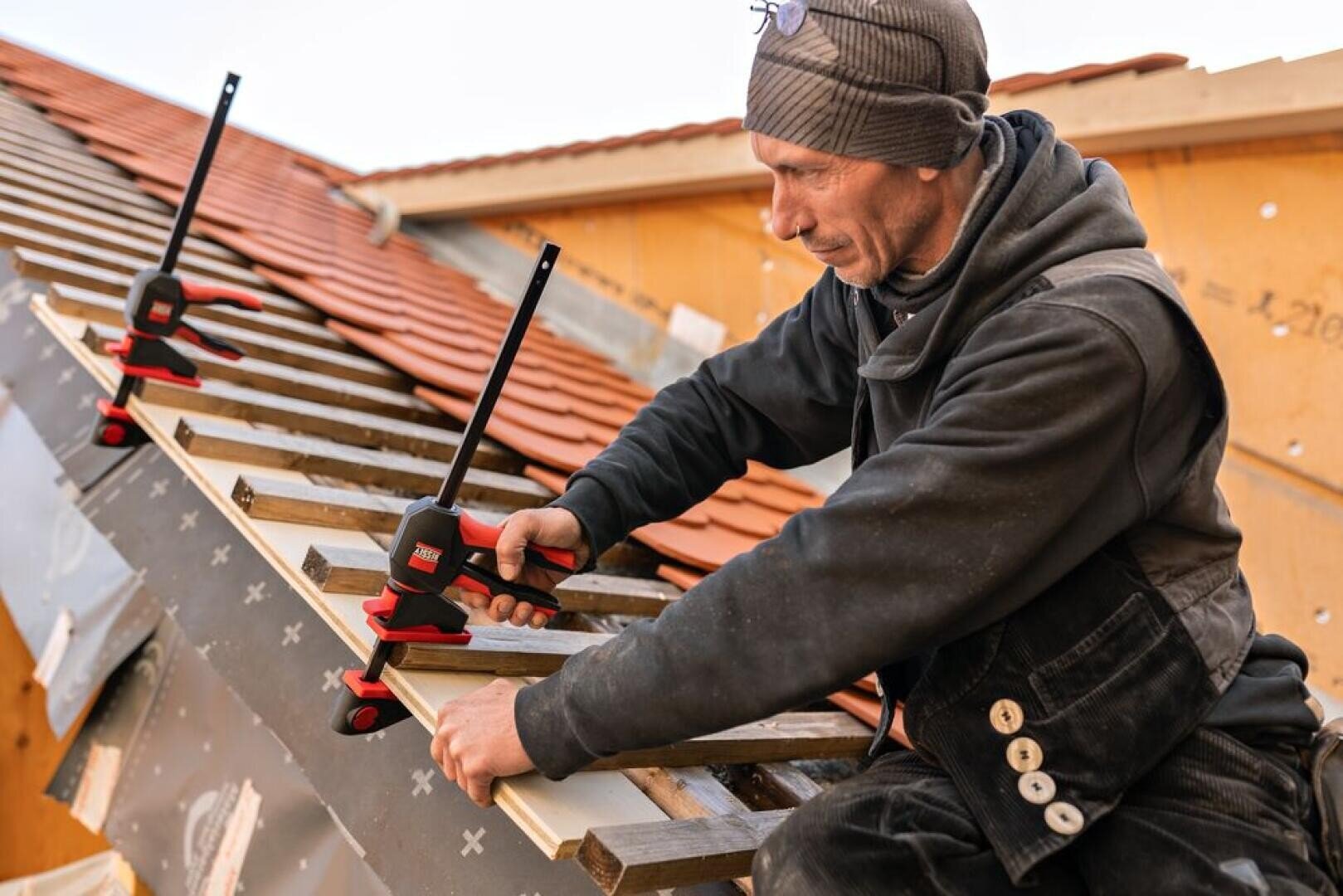 A worker in a cap and dungarees fastens wooden planks with clamps to a sloping roof under construction, with red roof tiles and wooden beams visible in the background.