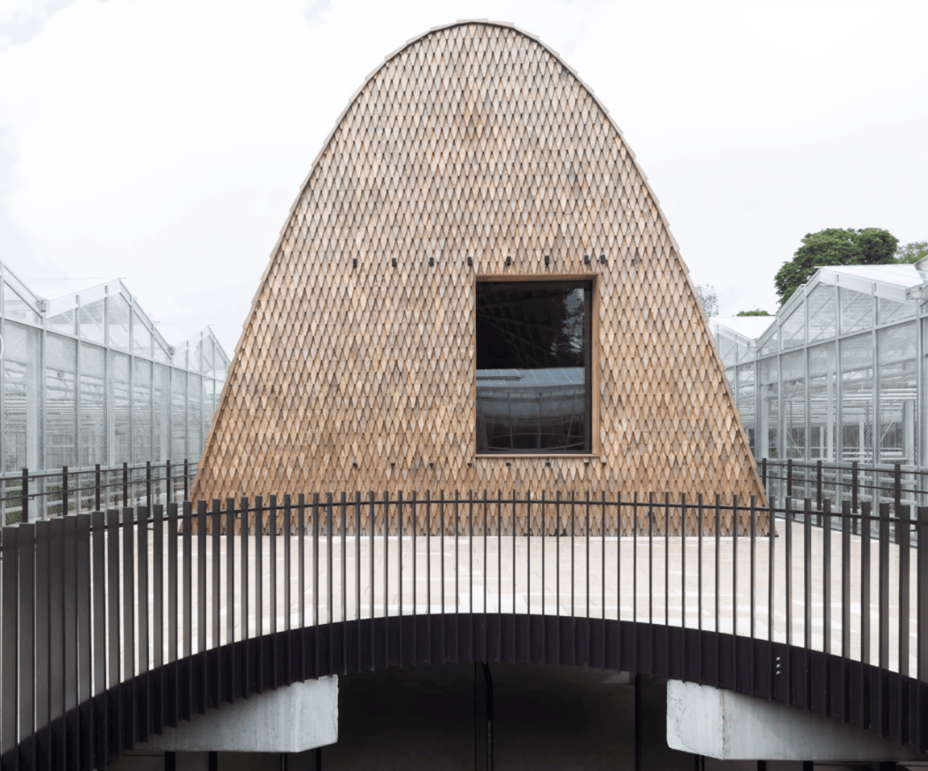 A dome-shaped building with a textured, woven exterior and a single square window. A curved black metal railing leads up to the entrance. Glass structures and green areas can be seen in the background.