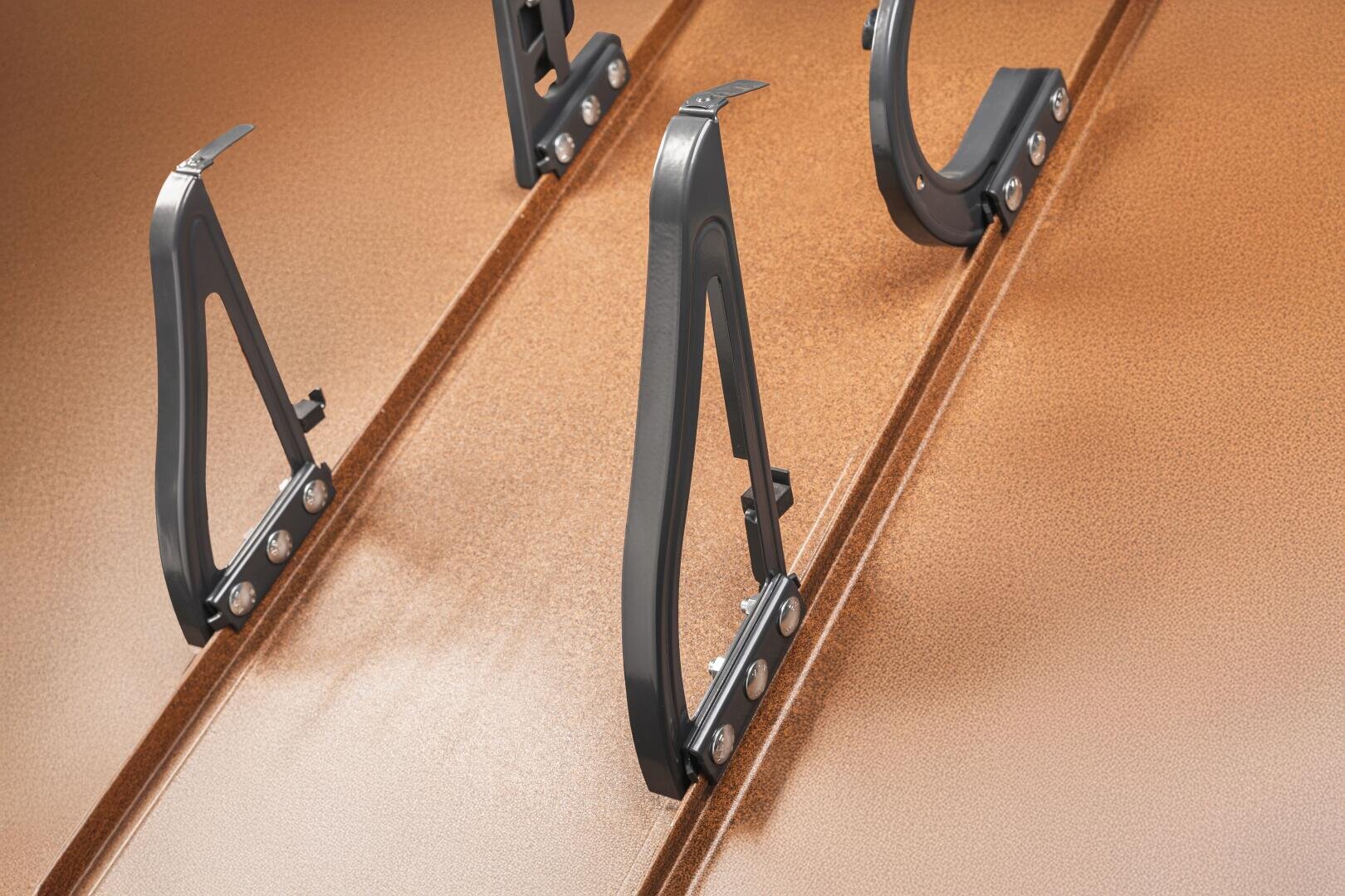 Nahaufnahme von schwarzen Metallklammern, die auf einer geriffelten, kupferfarbenen Metallfläche montiert sind, wahrscheinlich Teil eines Überdachungssystems zur Befestigung von Geräten oder Platten.