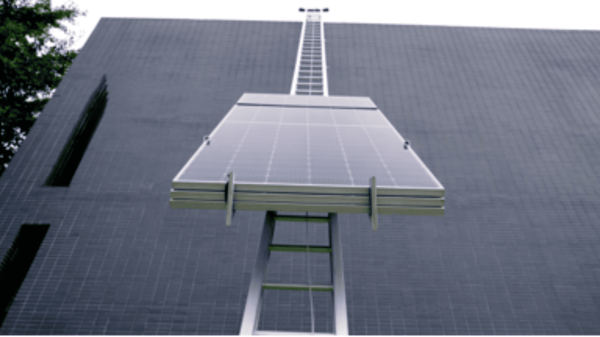 Large solar panel mounted on a metal structure is attached to the side of a modern, dark gray brick building, viewed from below with part of a tree visible on the left.