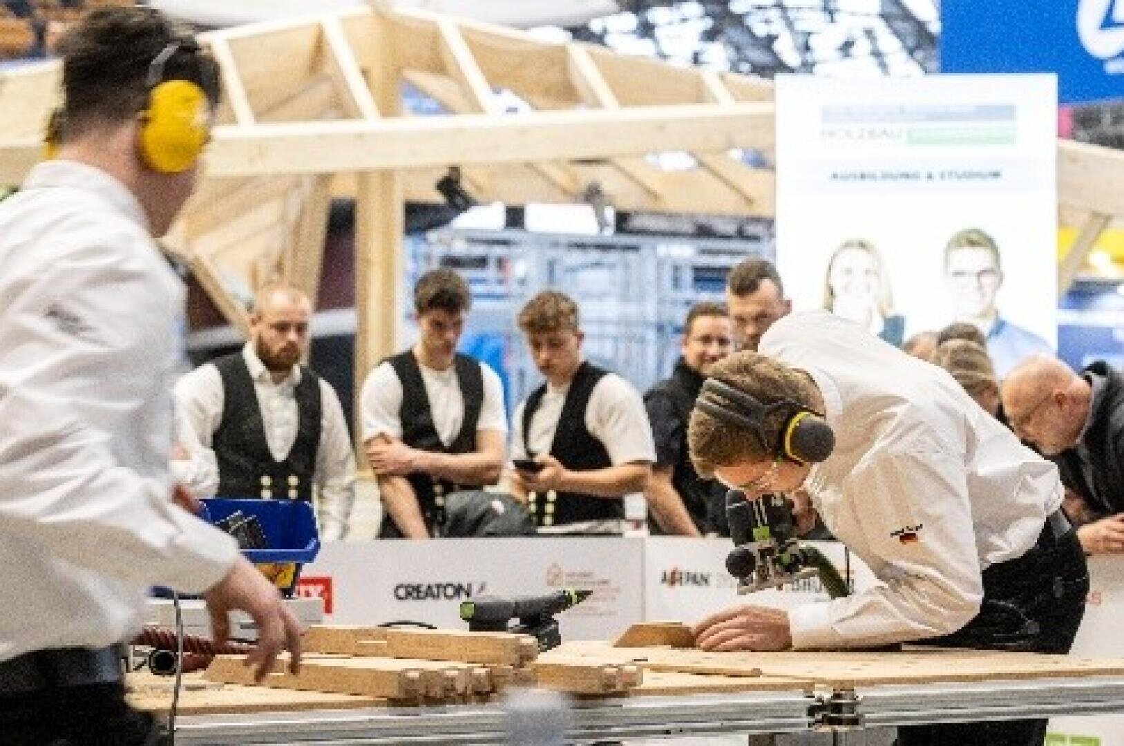 Ein Mann mit Sicherheitskopfhörern bearbeitet ein Stück Holz mit einem Elektrowerkzeug, während andere in einer Werkstatt oder bei einem Wettbewerb genau zusehen. Im Hintergrund stehen mehrere Schaulustige, einige in Uniform.