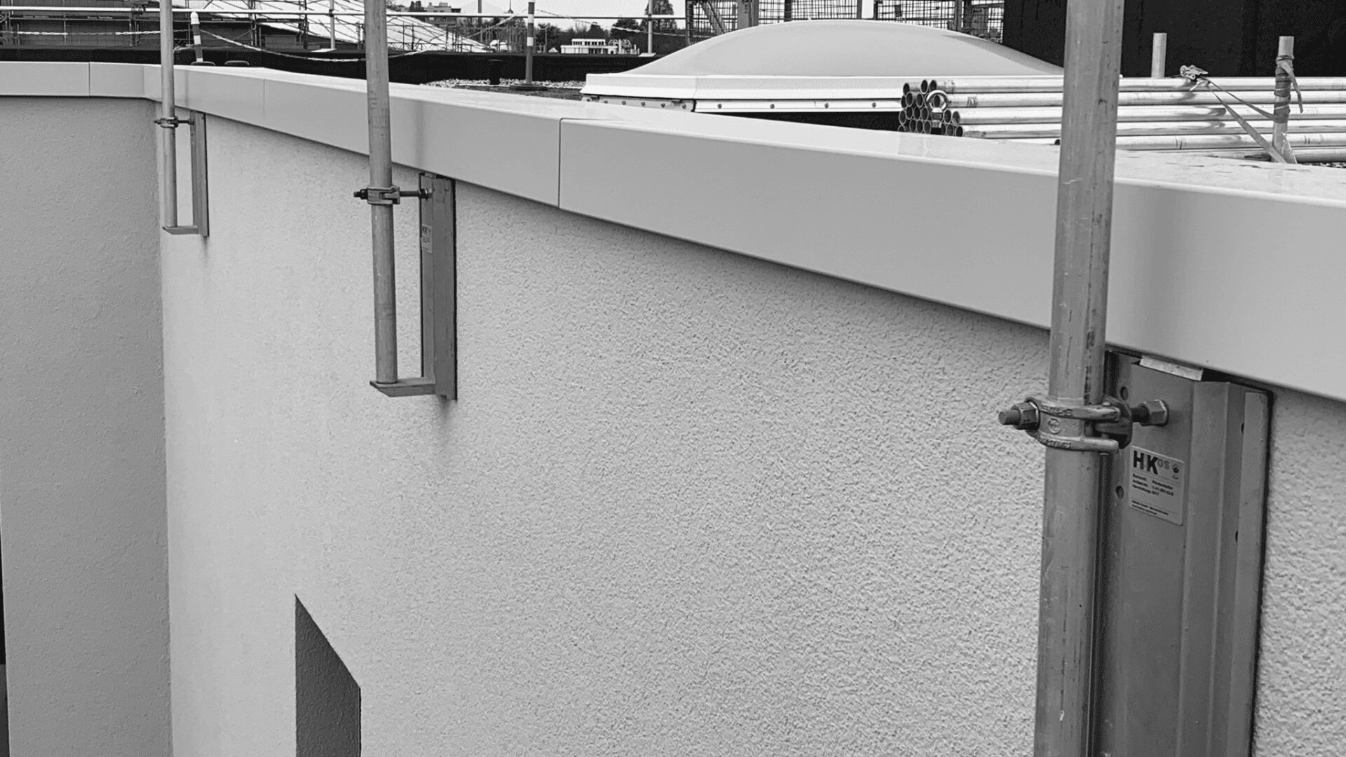 Schwarz-Weiß-Bild einer Gebäudewand mit provisorisch angebrachten Metallgerüststützen. Die Wand ist glatt und hat ein kleines Fenster. Im Hintergrund sind Baumaterialien auf dem Dach sichtbar.