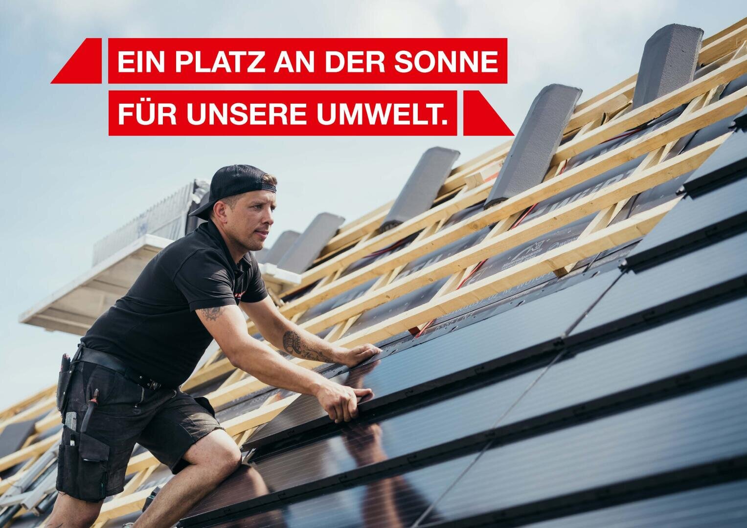 Ein Mann installiert Solarzellen auf einem Schrägdach unter einem teilweise bewölkten Himmel. Auf roten Bannern steht der deutsche Text: EIN PLATZ AN DER SONNE FÜR UNSERE UMWELT.
