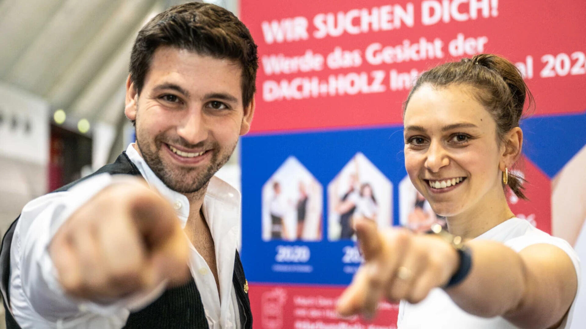 Ein lächelnder Mann und eine lächelnde Frau zeigen in die Kamera. Sie stehen vor einem rot-blauen Schild mit dem deutschen Text „WIR SUCHEN DICH!“ und weiteren Informationen zu einer Veranstaltung.