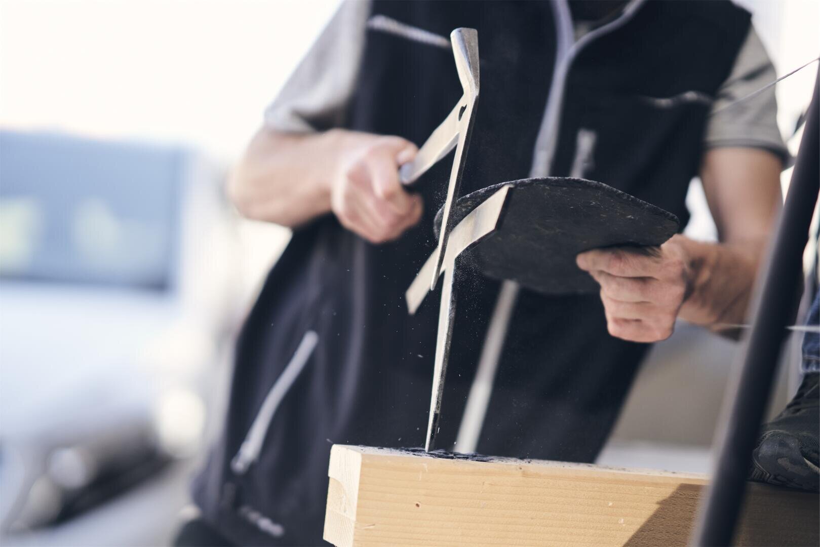 Eine Person sägt mit einer Handsäge durch ein Stück Material, wobei Sägespäne aufgewirbelt werden. Die Person trägt eine Weste und ein kurzärmeliges Hemd, und die Arbeit wird auf einer Holzoberfläche ausgeführt.