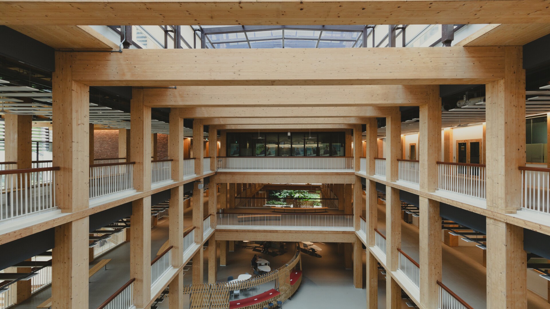 Offenes, mehrstöckiges Atrium in einem modernen Gebäude mit freiliegenden Holzbalken, Glasdecke und Innenbalkonen, umgeben von weißen Geländern, das ein helles und geräumiges architektonisches Design zur Schau stellt.
