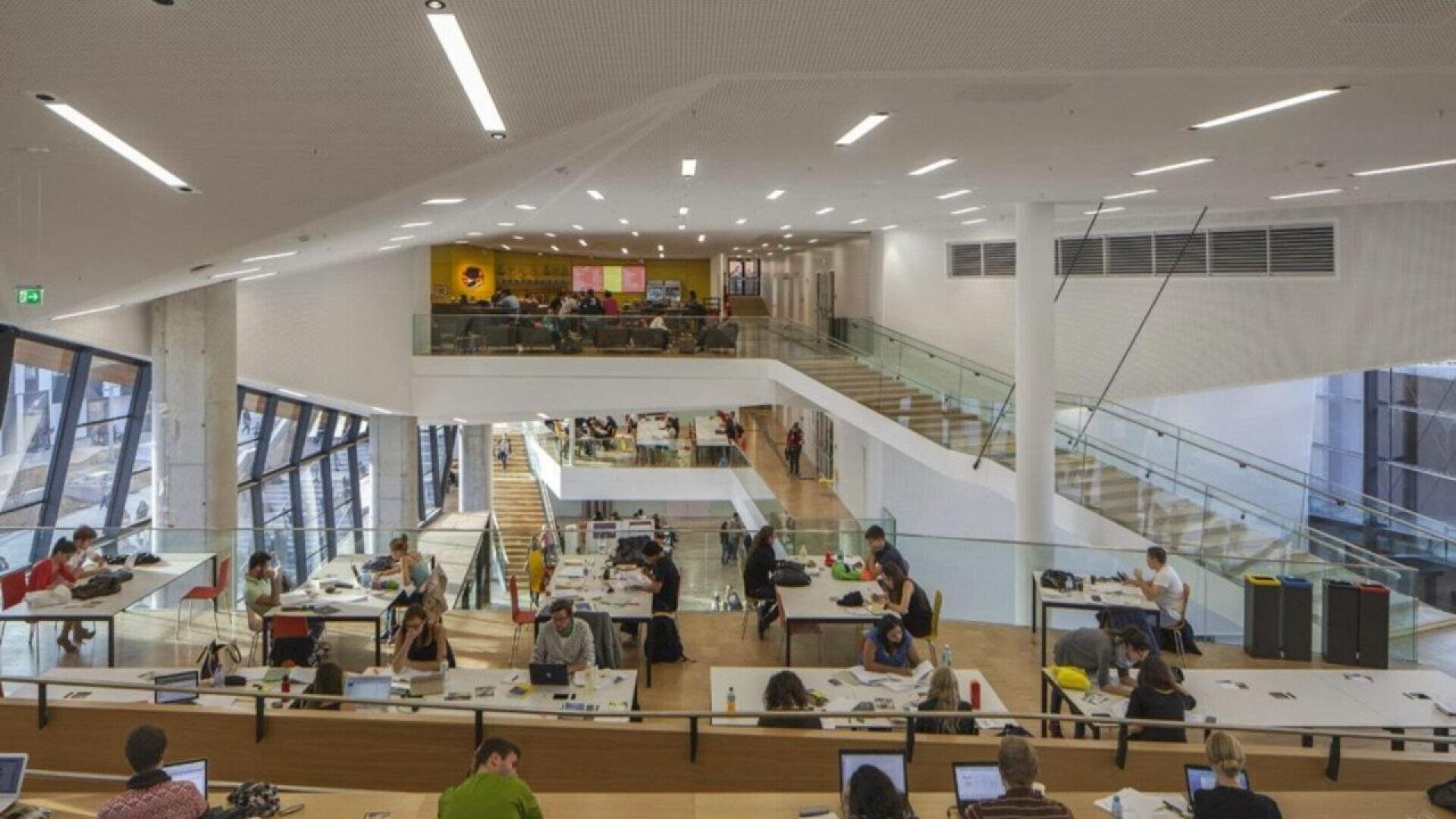 A spacious, modern library with large windows, multiple levels, and people studying at long desks. Some individuals work on laptops, while others read or write. The atmosphere appears quiet and focused.