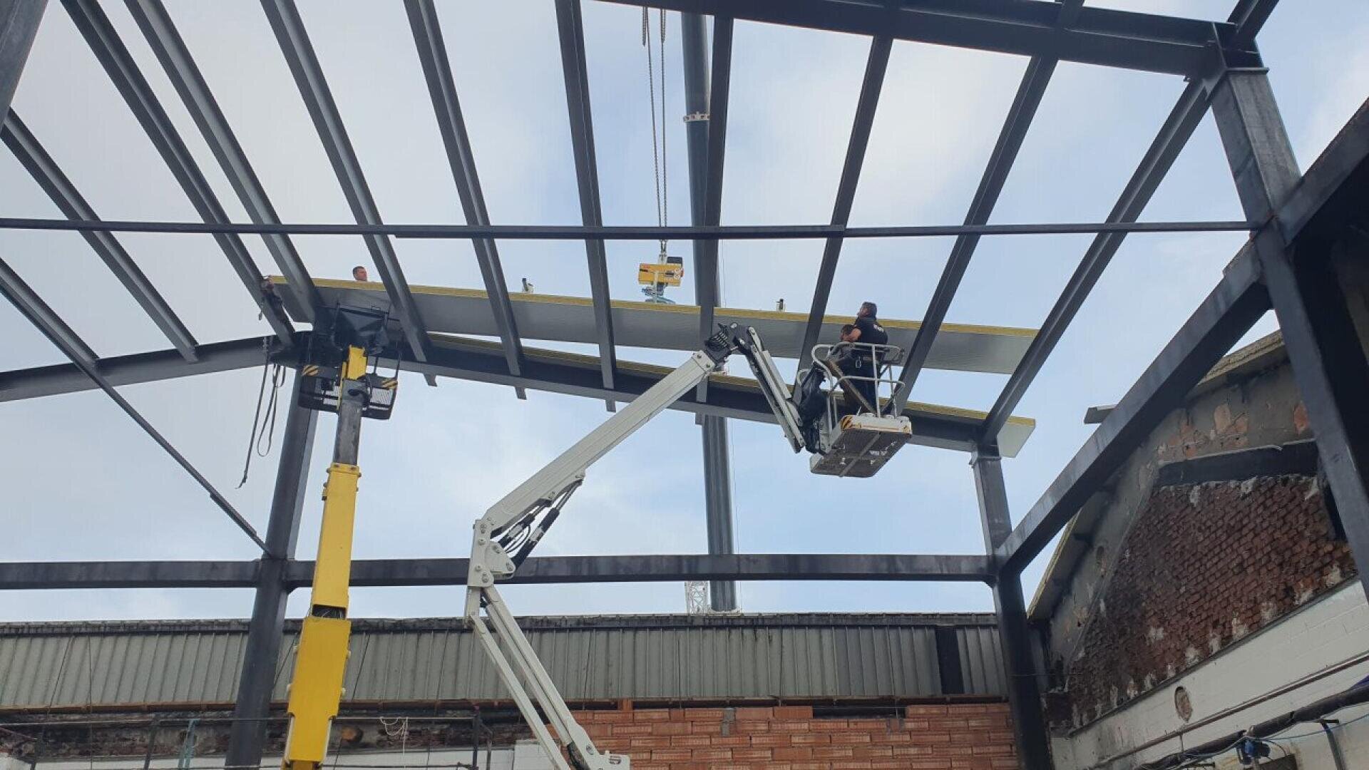 Bauarbeiter montieren auf einer Hebebühne Metallträger an einem Stahlgerüst eines Gebäudes, wobei ihnen über ihnen unter einem teilweise bewölkten Himmel ein Kran hilft.