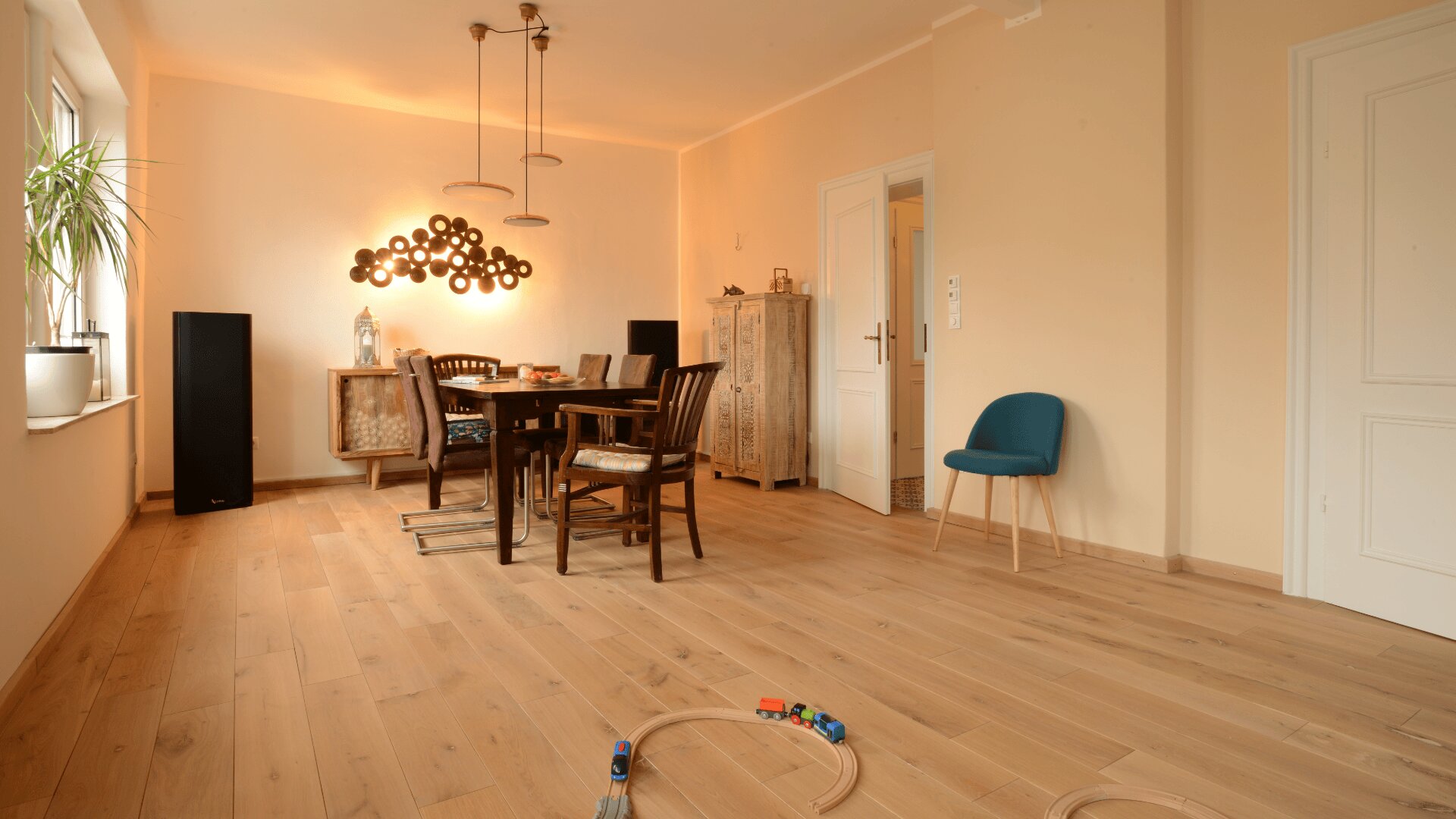 Ein Esszimmer mit Holzboden, einem Tisch mit Stühlen, dekorativer Beleuchtung, einem blauen Stuhl an der Wand und einer Spielzeugeisenbahn auf dem Boden. Sonnenlicht fällt durch ein Fenster mit grünen Pflanzen auf der Fensterbank.