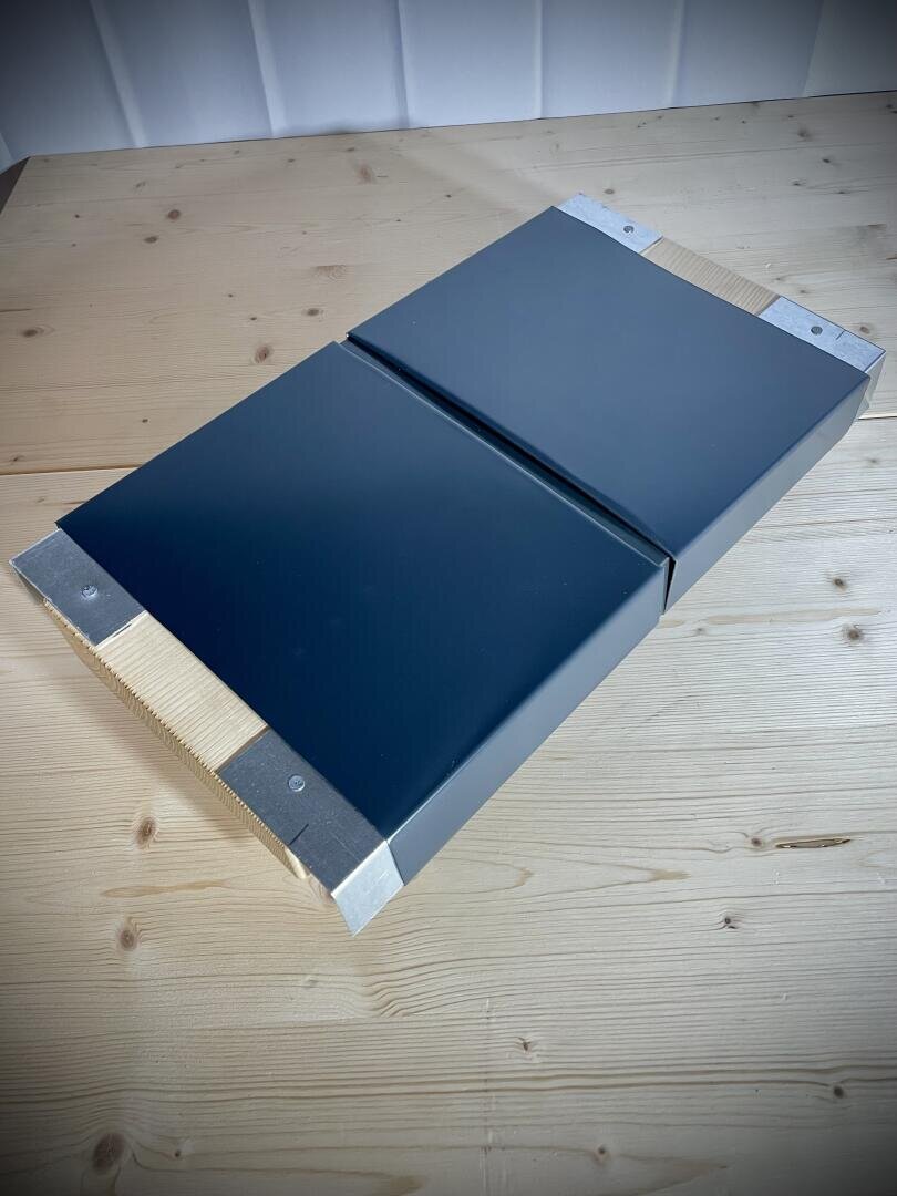 Zwei schwarze, rechteckige Metallplatten mit silbernen Enden liegen nebeneinander auf einer hellen Holzoberfläche und bilden eine symmetrische Form. Jede Platte scheint an den Enden einen Holzblock darunter zu haben.
