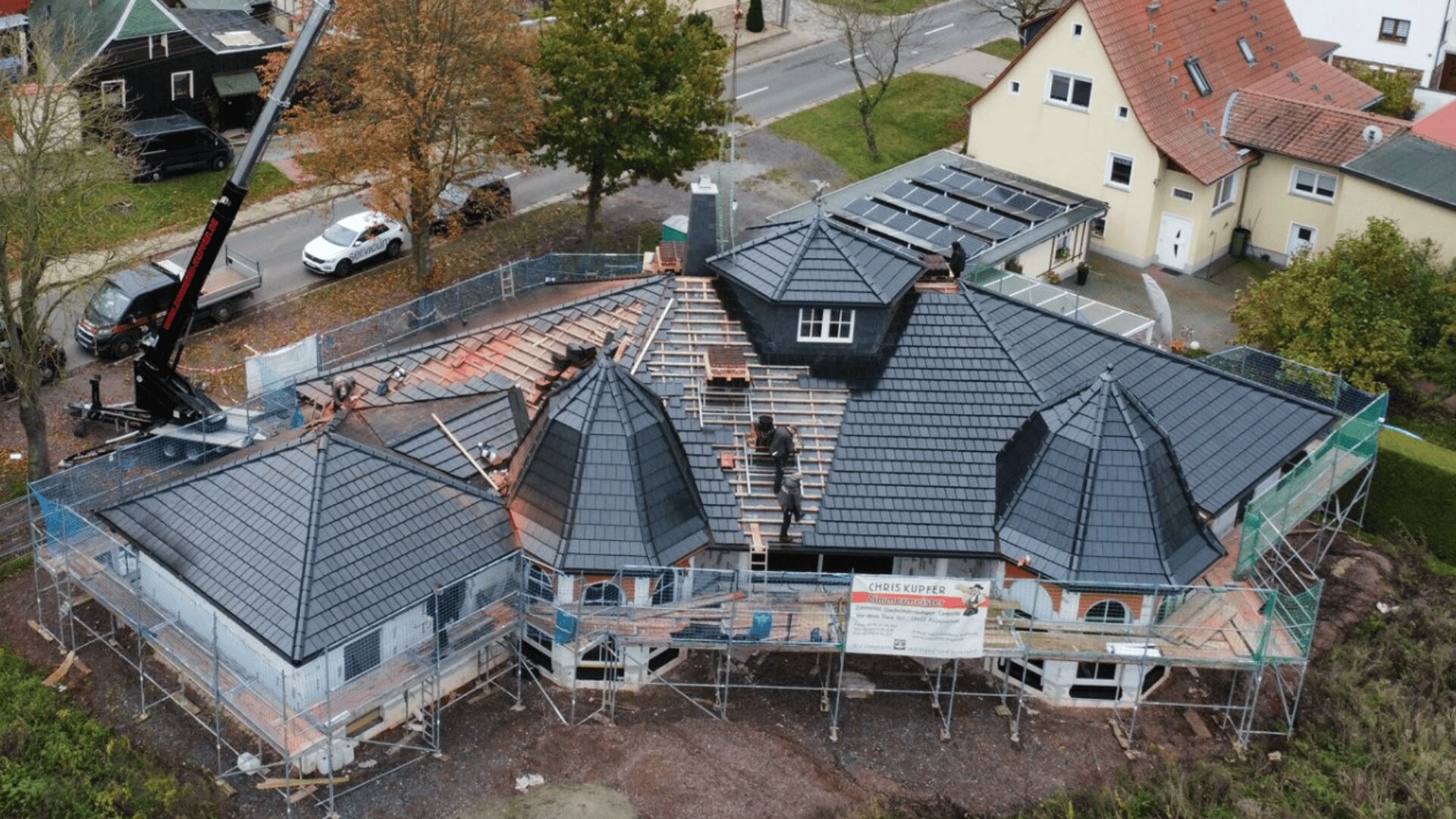 Luftaufnahme eines im Bau befindlichen Hauses mit Gerüsten; Arbeiter verlegen dunkle Dachziegel, ein Kran steht in der Nähe. Um das Haus herum befinden sich Bäume, Straßen und Nachbarhäuser.