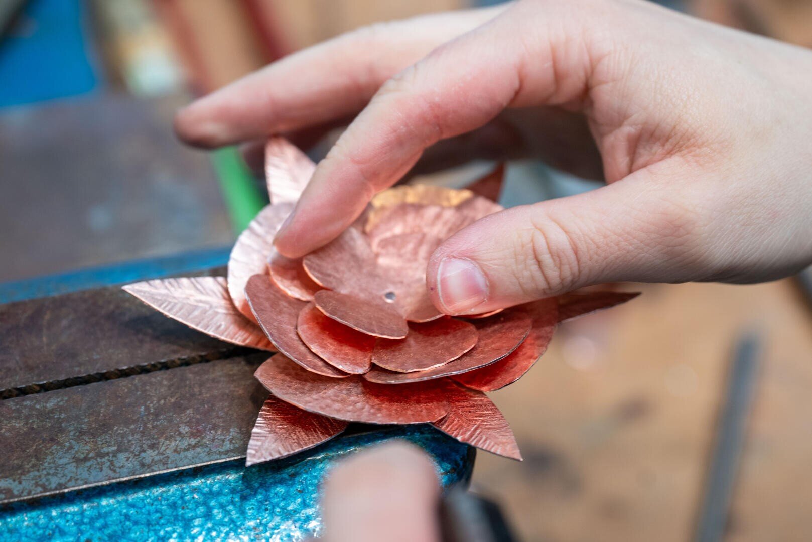 Die Hände einer Person fügen geschichtete Kupferblütenblätter in Form einer Blume zusammen, wobei ein Metallschraubstock auf einer Werkbank als Stütze dient.
