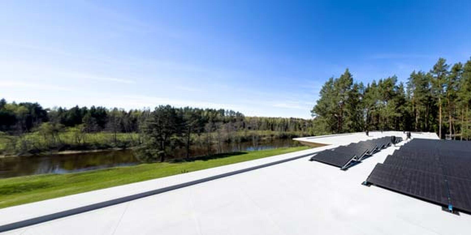 A row of solar panels is located on a flat roof overlooking a river, surrounded by green grass and dense forest under a clear blue sky.