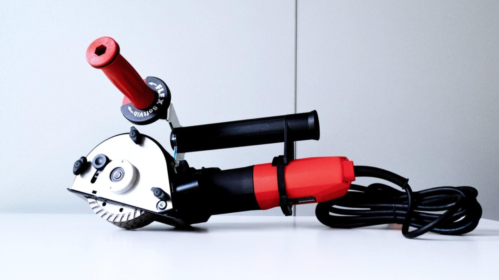 A red and black handheld angle grinder with two handles and a coiled power cord rests on a white surface against a plain light background.