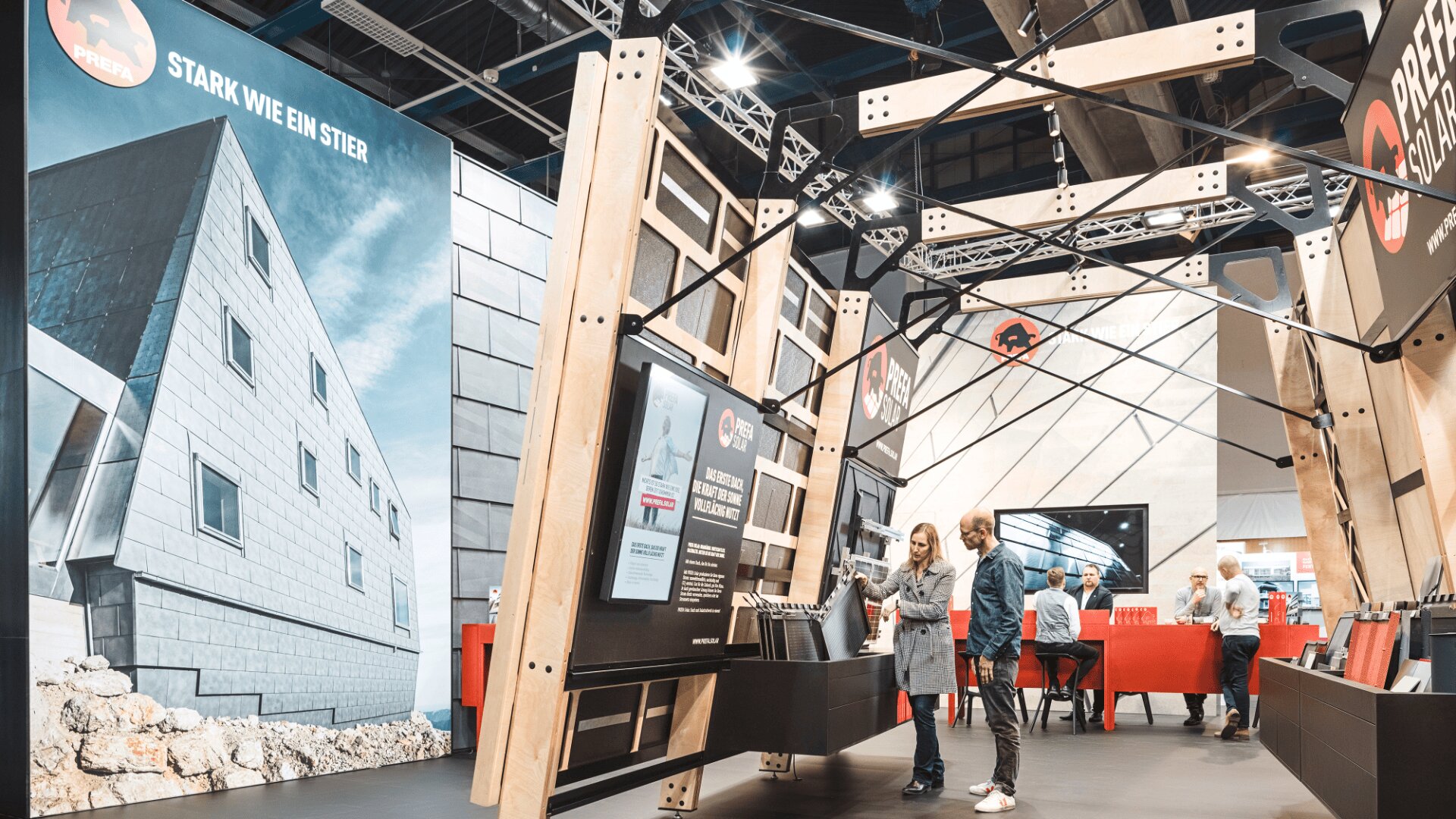 Menschen interagieren mit Displays an einem modernen Messestand mit großen Bildern einzigartiger Gebäude, hölzernen Strukturelementen und Bannern mit deutschem Text. Heller, offener Raum mit einigen Personen im Hintergrund.