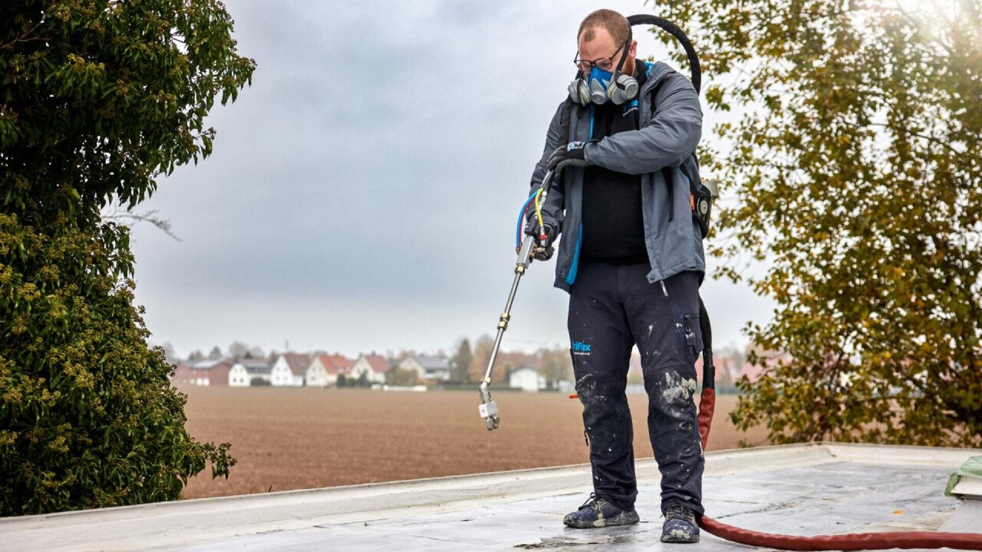 A worker wearing a respirator mask and work clothes stands on a flat roof, holding spraying equipment. The background shows a field, trees, and houses under a cloudy sky.