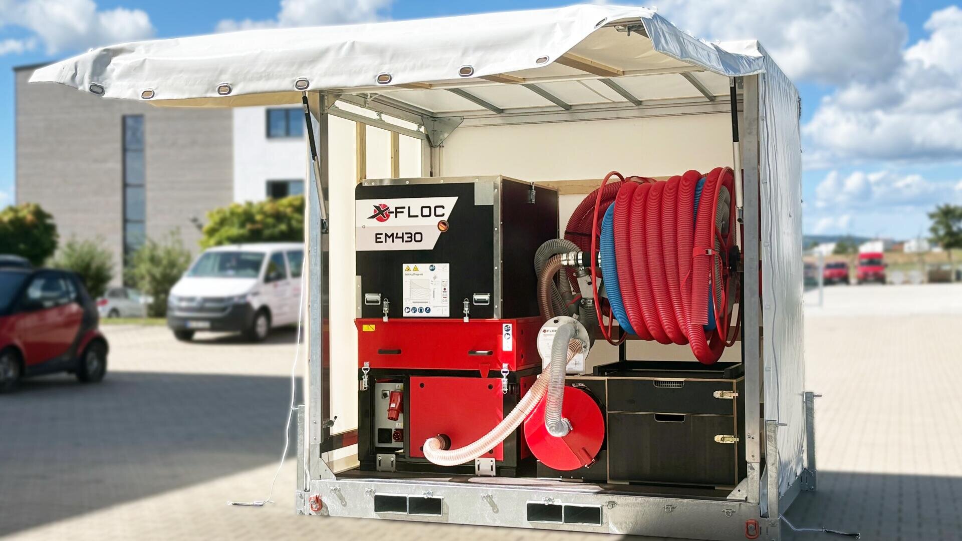 Eine X-Floc Einblasmaschine EM430 ist in einem offenen Anhänger mit einer Schlauchtrommel und roten Schläuchen montiert. Die Plattform steht auf einer gepflasterten Fläche mit Gebäuden und Fahrzeugen im Hintergrund.