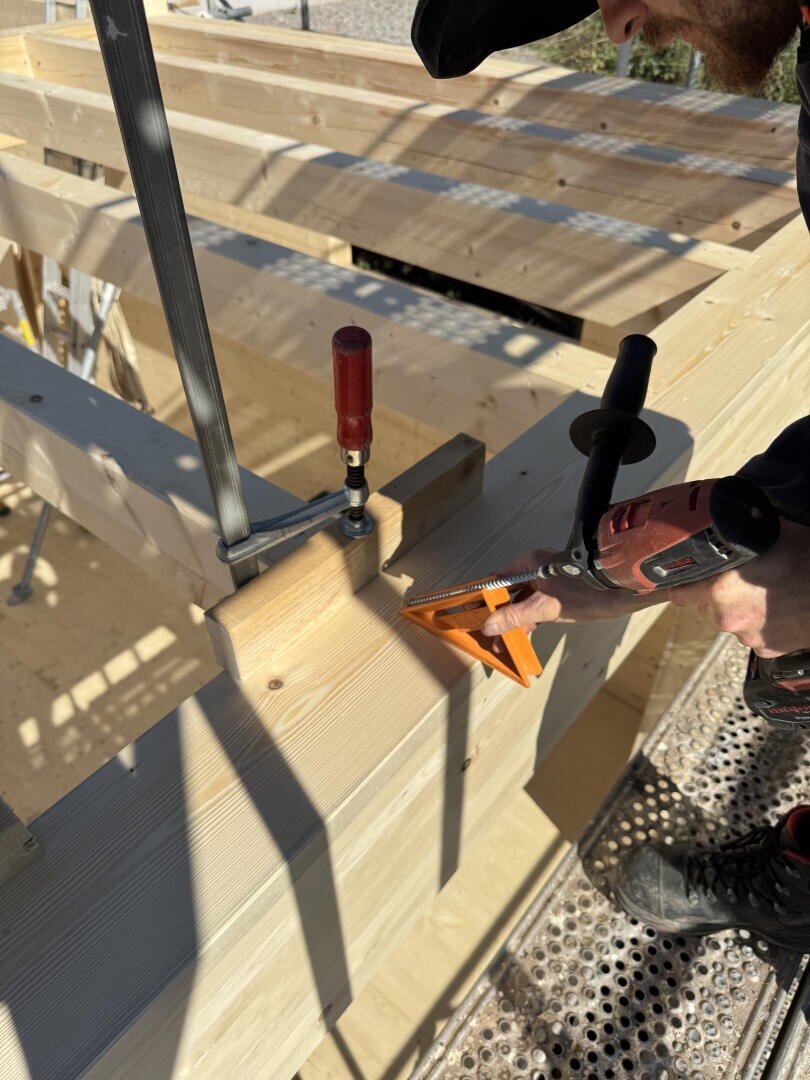 Eine Person arbeitet mit einer Bohrmaschine und einem orangefarbenen Zimmermannswinkel an einer Holzkonstruktion und befestigt Balken mit Zwingen auf einer Baustelle.