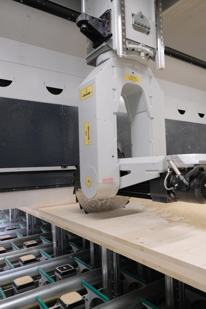 Eine Nahaufnahme einer industriellen Sägemaschine, die ein Holzbrett durchschneidet. Auf der Oberfläche sammelt sich Sägemehl, und rund um den Schneidebereich sind mechanische Komponenten sichtbar.