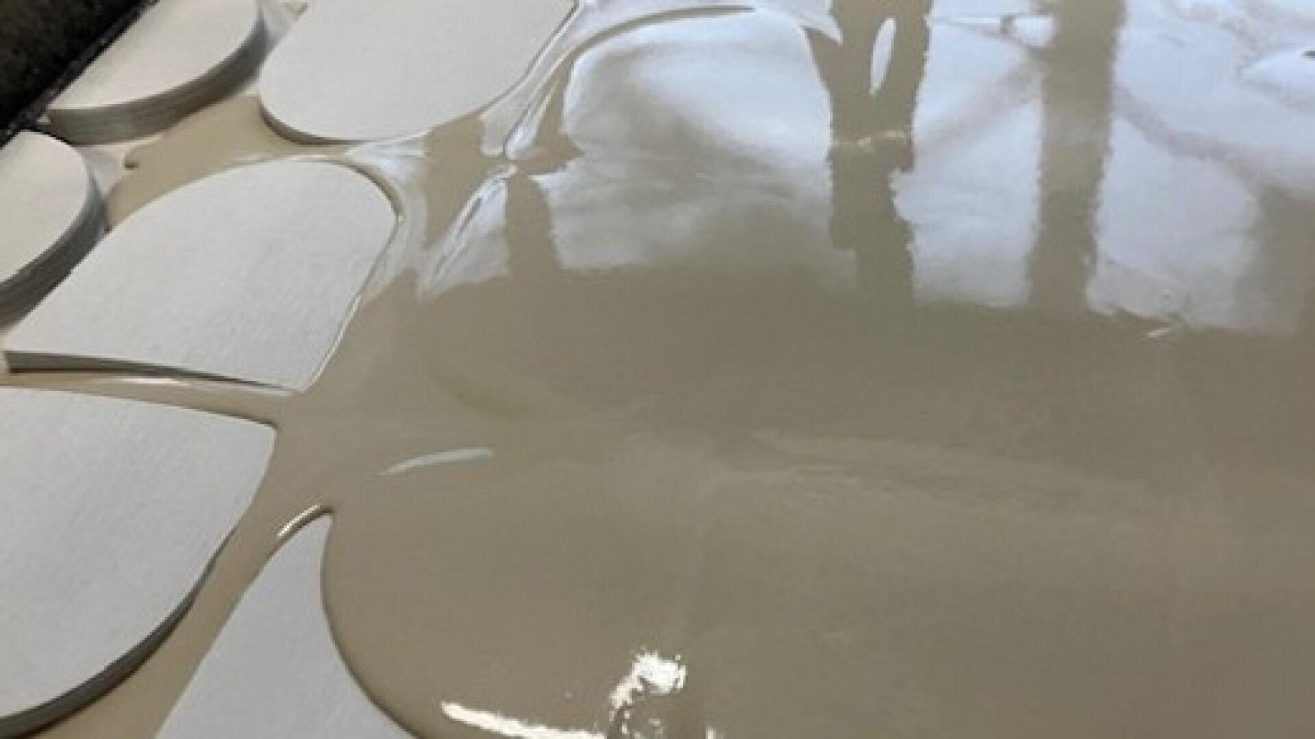 Eine Nahaufnahme einer glatten, glänzenden beigen Flüssigkeit, die über kreisförmige weiße Formen gegossen wird, möglicherweise in einer Produktions- oder Lebensmittelzubereitungsumgebung. Auf der Oberfläche der Flüssigkeit sind Reflektionen zu sehen.