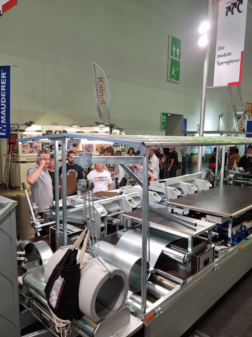 Eine Gruppe von Menschen beobachtet auf einer Messe oder Ausstellung eine große Industriemaschine, die Bleche verarbeitet. Im Hintergrund sind Banner, Sicherheitsschilder und verschiedene Stände zu sehen.