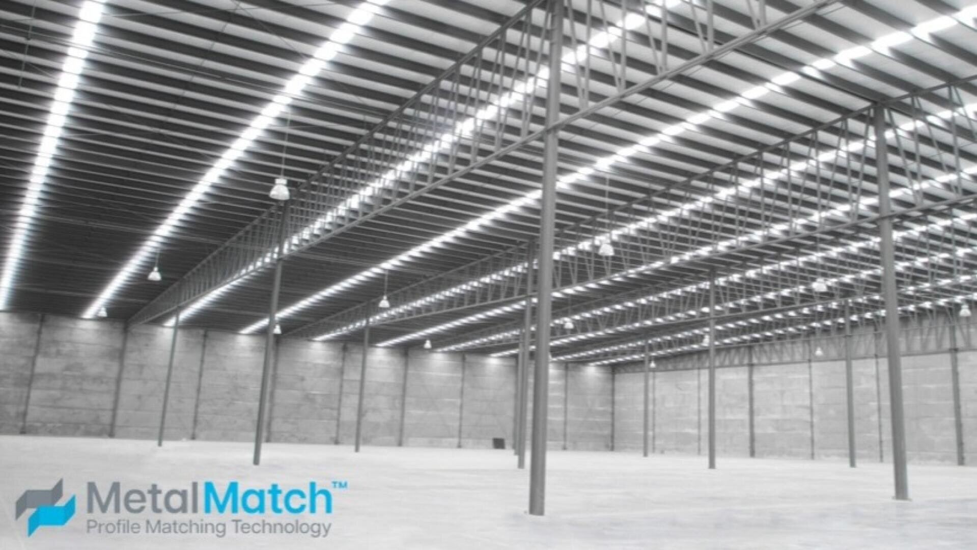 A large, empty warehouse with high ceilings, metal beams and bright overhead lighting. The floor is concrete and the space is open and spacious. The MetalMatch logo appears in the bottom left corner.