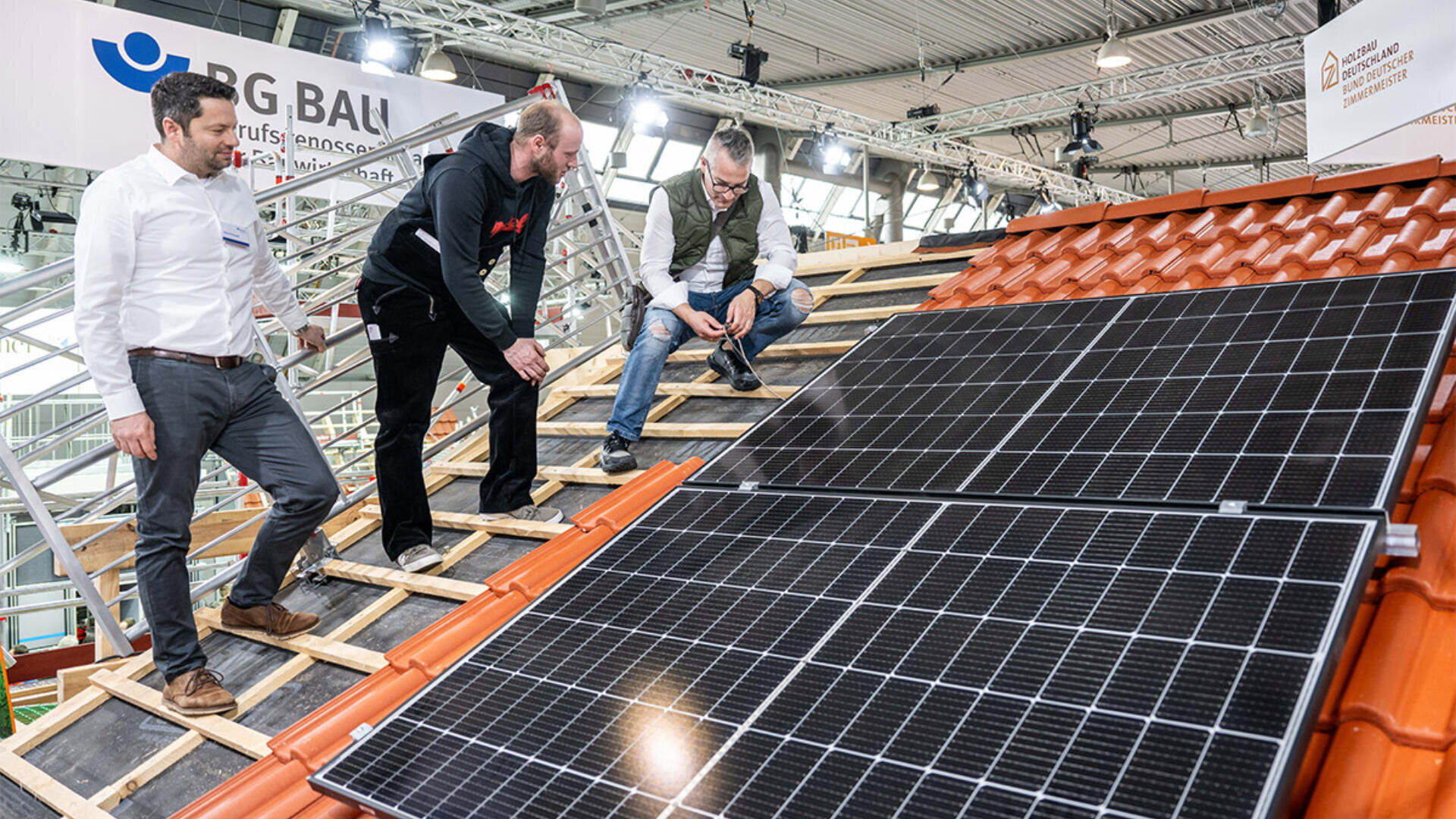 Drei Männer untersuchen und diskutieren die Installation von Solarmodulen auf einem roten Ziegeldach in einer Ausstellungshalle. Im Hintergrund sind Gerüste und Beschilderungen zu sehen.