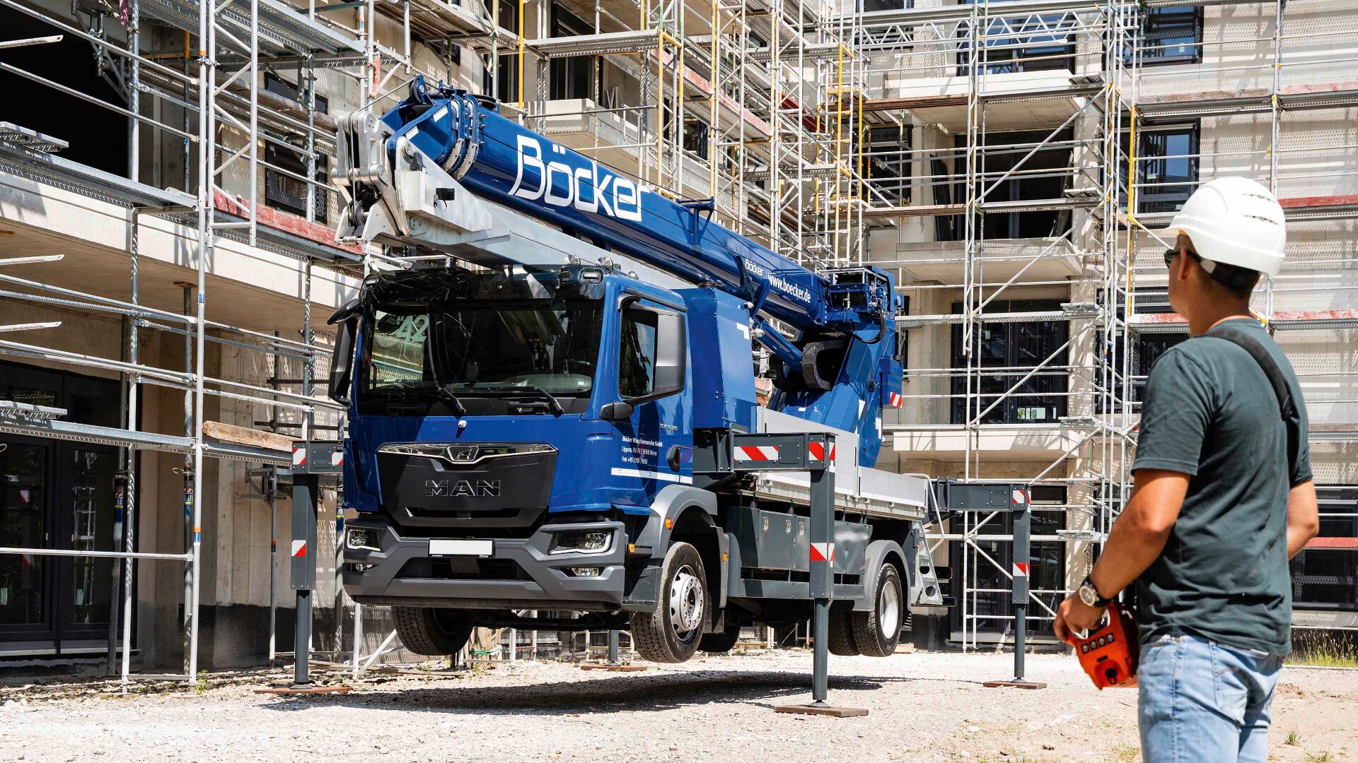 Ein blauer Bauwagen mit einem Kranaufsatz wird an seinen Stützbeinen in der Nähe eines im Bau befindlichen Gebäudes vom Boden abgehoben; ein Arbeiter mit weißem Schutzhelm steht in der Nähe und hält eine Fernsteuerung.
