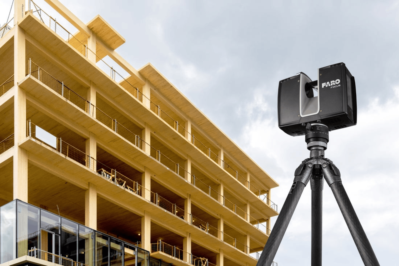 Ein FARO 3D-Laserscanner auf einem Stativ steht vor einem im Bau befindlichen mehrstöckigen, modernen Holzgebäude vor einem bewölkten Himmel.