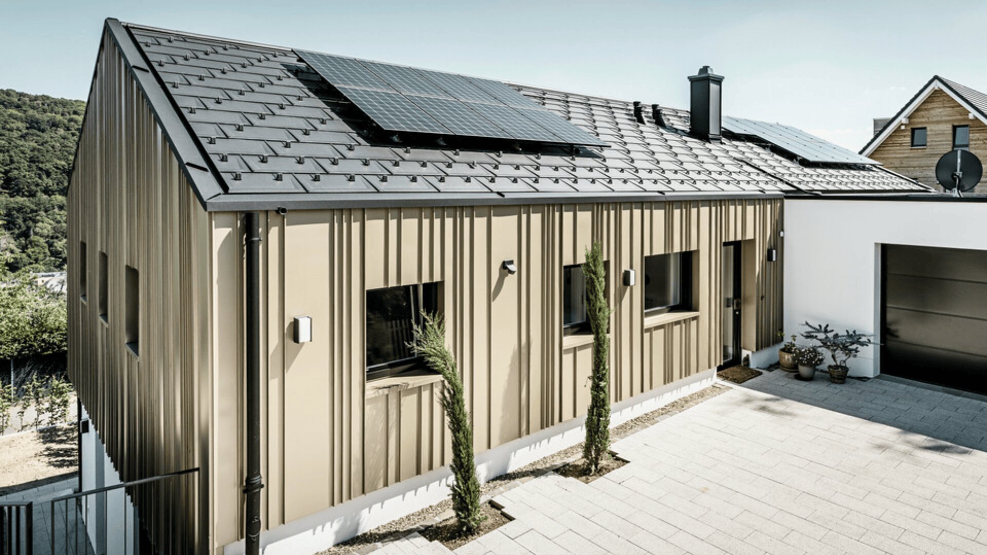 Modernes Haus mit Metalldach, Solarmodulen, beiger vertikaler Fassade und mehreren kleinen rechteckigen Fenstern. In der gepflasterten Einfahrt neben dem Haus stehen junge Bäume und Topfpflanzen.