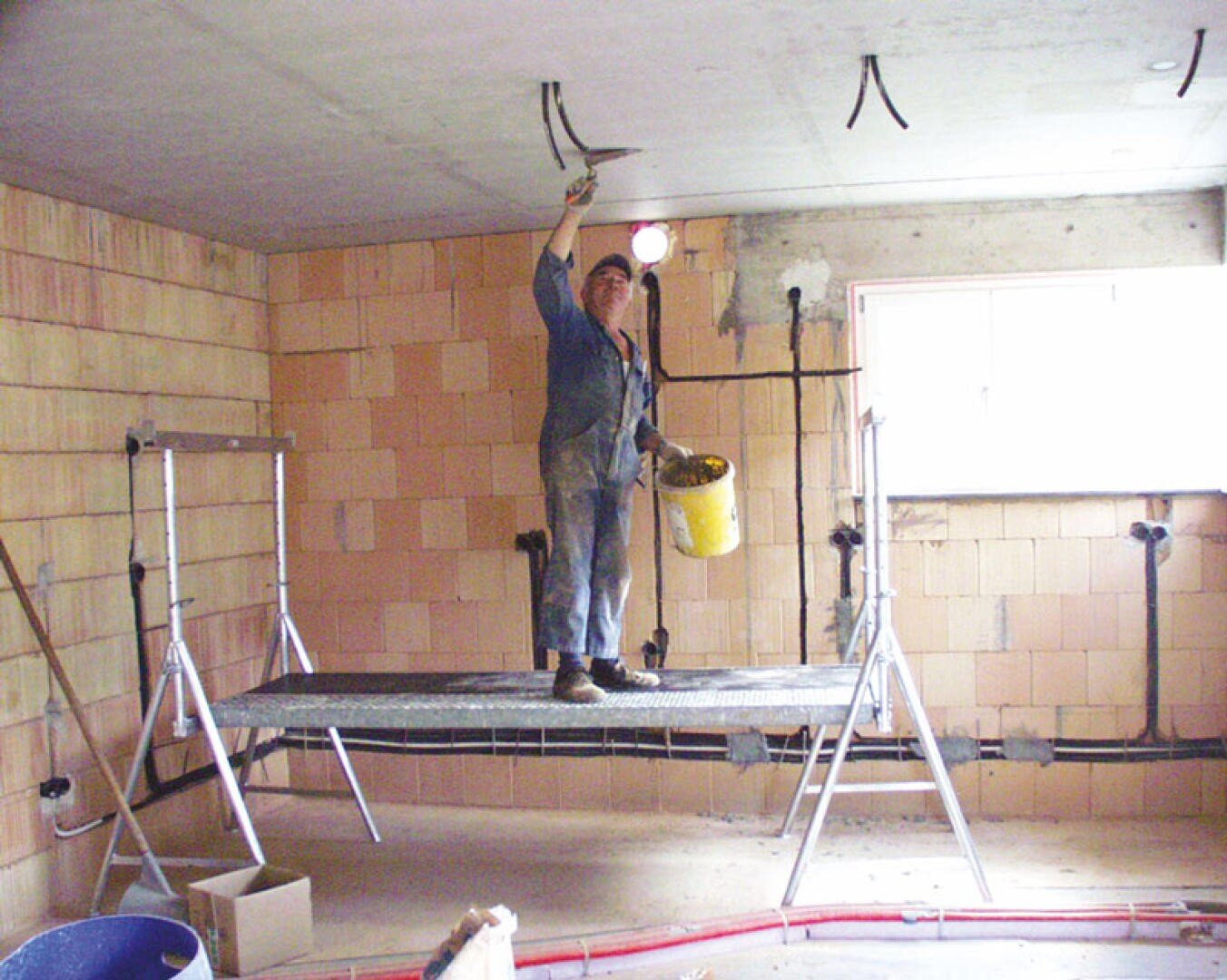 A construction worker stands on a platform, holding a trowel in one hand and a yellow bucket in the other, applying plaster to the ceiling of an unfinished room with exposed brick walls.