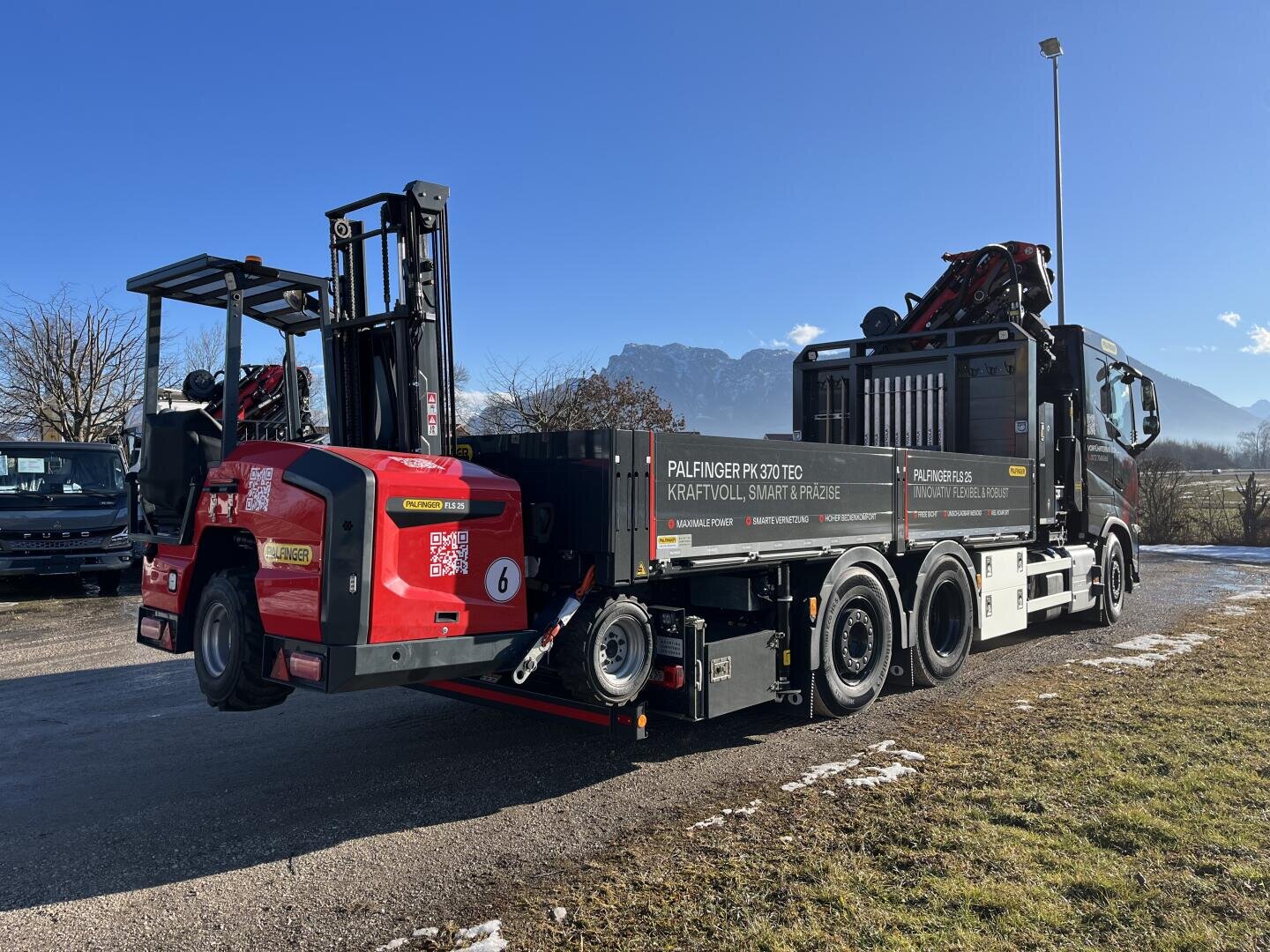Ein großer schwarzer Lastwagen mit einem Kran transportiert einen roten Gabelstapler auf seiner Ladefläche. Die Szene spielt im Freien, mit einem klaren blauen Himmel, Bergen im Hintergrund und einigen Schneeflecken auf dem Boden.