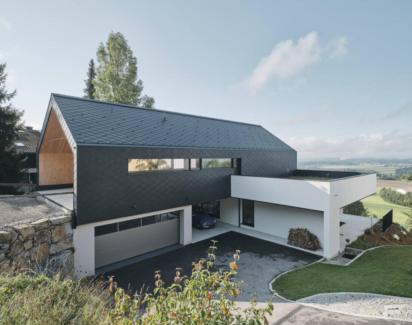 A modern house with a black pitched roof, white and dark geometric exterior walls, a large garage and a driveway, standing on a green hill with open sky in the background.
