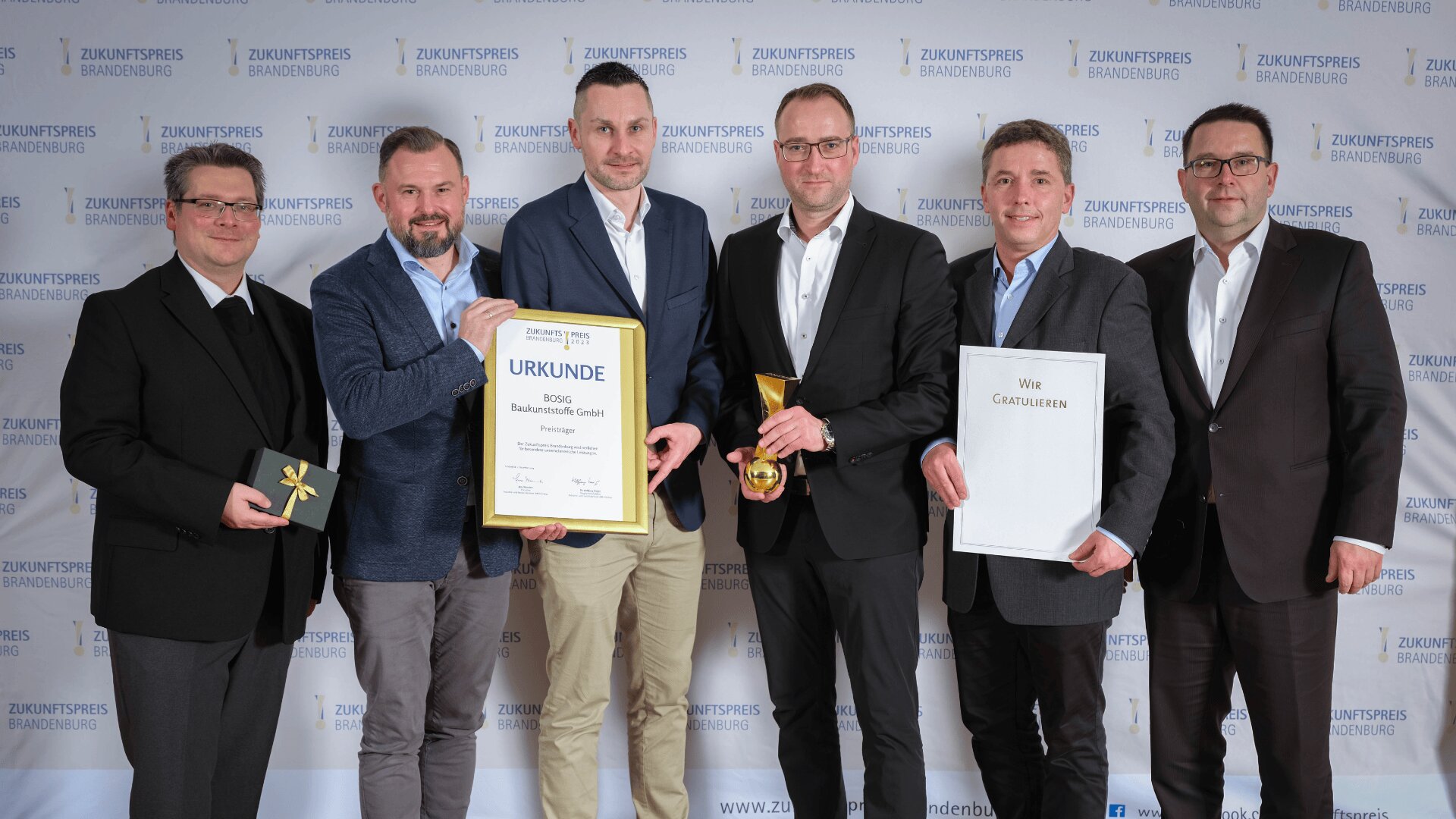 Sechs Männer in Anzügen stehen lächelnd zusammen und halten eine Urkunde, eine Trophäe, ein Glückwunschschreiben und eine kleine Geschenkbox vor einer Kulisse mit der Aufschrift „Zukunftspreis Brandenburg“ bei einer Preisverleihung in den Händen.