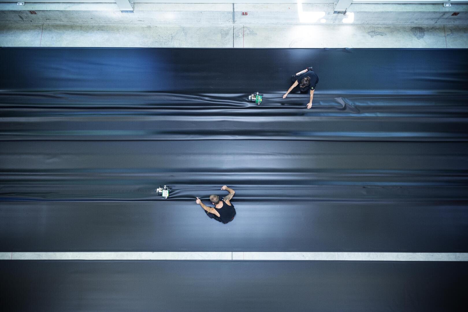 Two people work in an industrial environment on large, smooth black panels, pressing or smoothing the material with hand tools; the scene is seen from above.