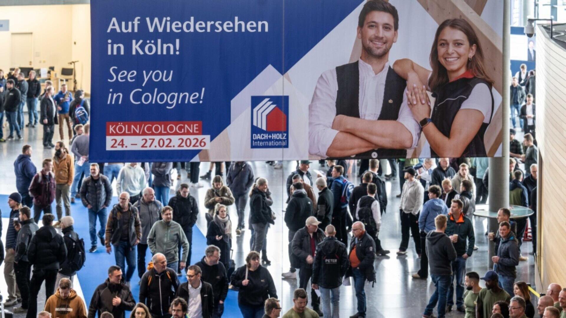 A large crowd walks in an indoor event hall under a banner that reads See you in Cologne! 24-27.02.2026 with two smiling people in work attire pictured on it. The banner also displays the DACH+HOLZ International logo.