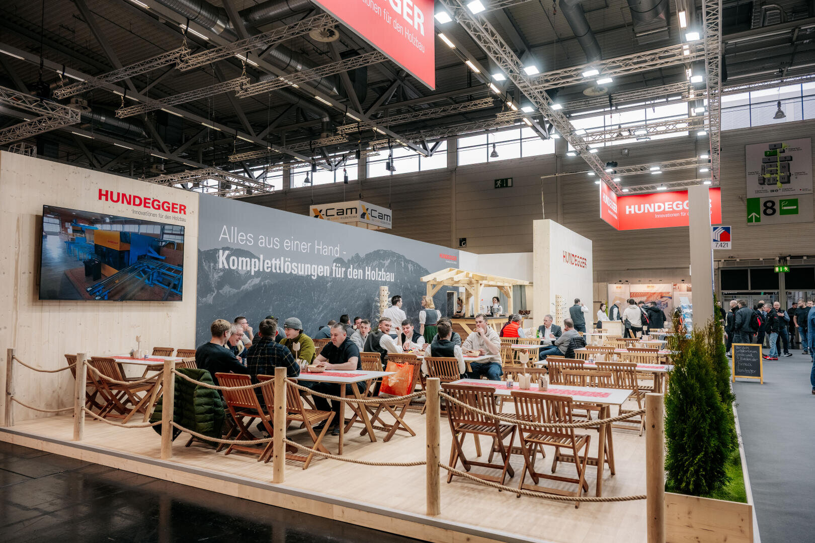 Ein Messestand mit Menschen, die an Holztischen sitzen, sich unterhalten und essen. Der Stand ist mit Holzdekor, einem großen Schild mit der Aufschrift Hundegger und einem Bildschirm mit Bildern ausgestattet. Im Hintergrund sind andere Stände und Besucher zu sehen.