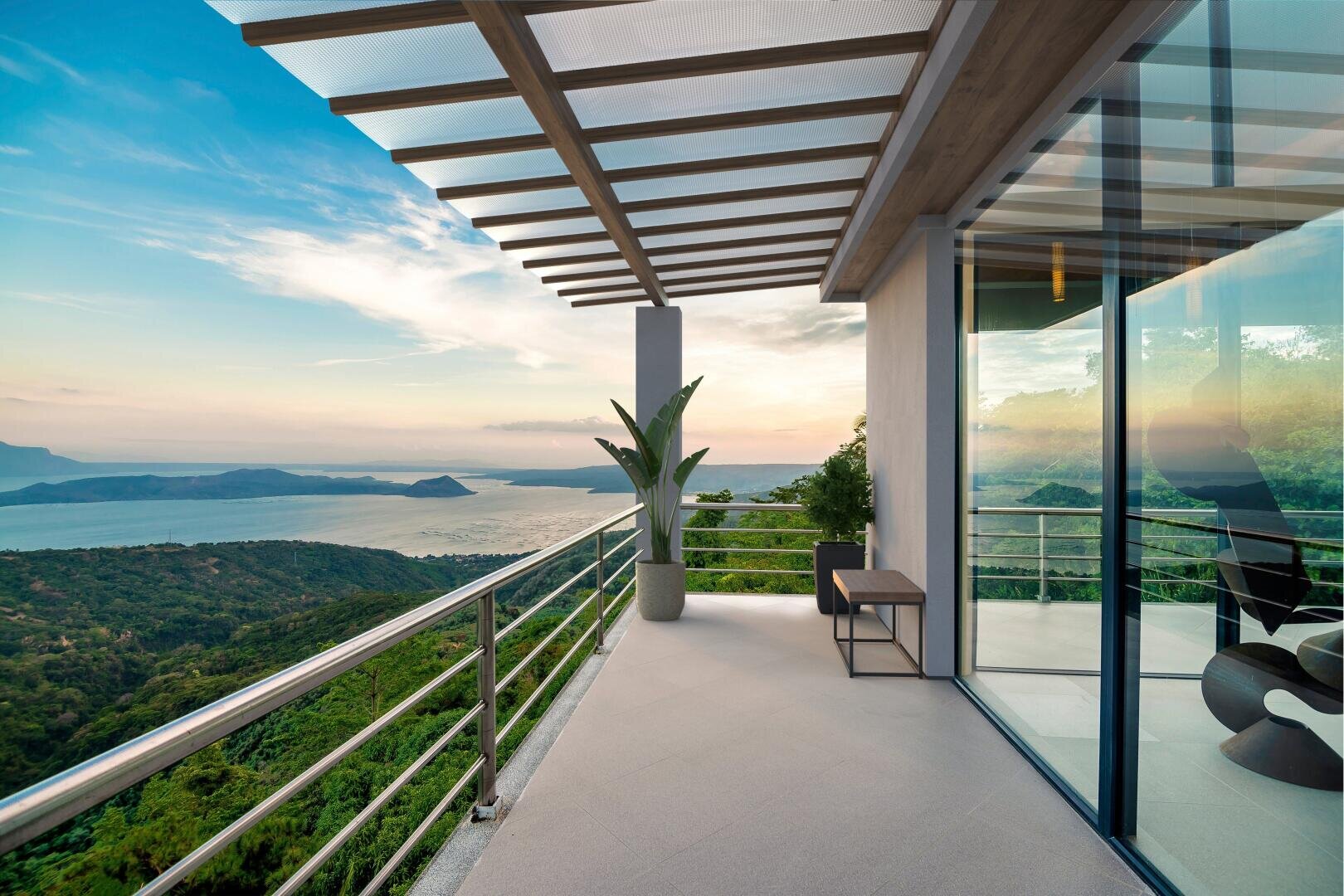 Moderner Balkon mit Glasgeländer und Pergoladach, mit Blick auf eine malerische Landschaft mit grünen Hügeln, einem großen Gewässer und fernen Bergen bei Sonnenuntergang; Topfpflanze und Gartenmöbel sichtbar.