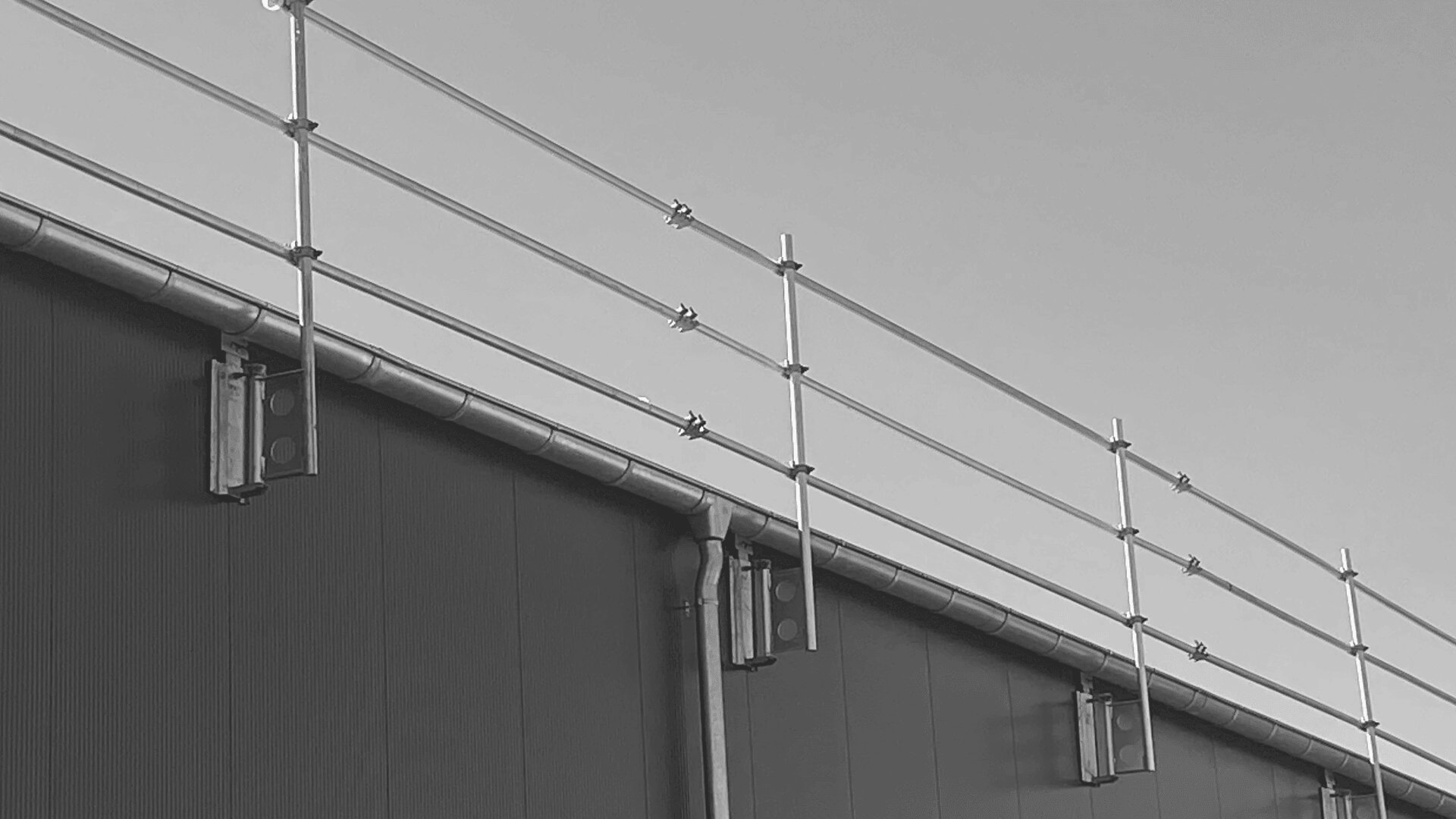 Ein Schwarzweißfoto eines Gebäudedachs mit metallenem Sicherheitsgeländer und Stützbalken, von unten vor einem klaren Himmel gesehen.