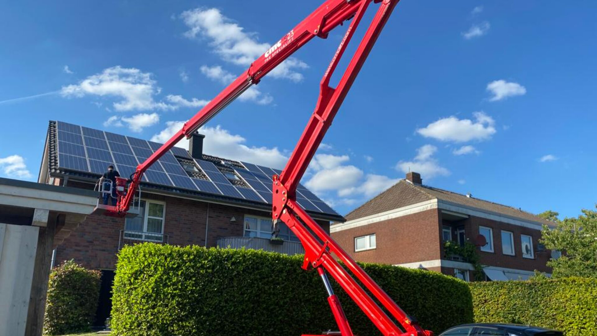 Unter einem blauen, bewölkten Himmel fährt eine rote Hebebühne auf das Dach eines Hauses mit Solarmodulen. Im Hintergrund sind ein zweites Haus und grüne Hecken zu sehen.