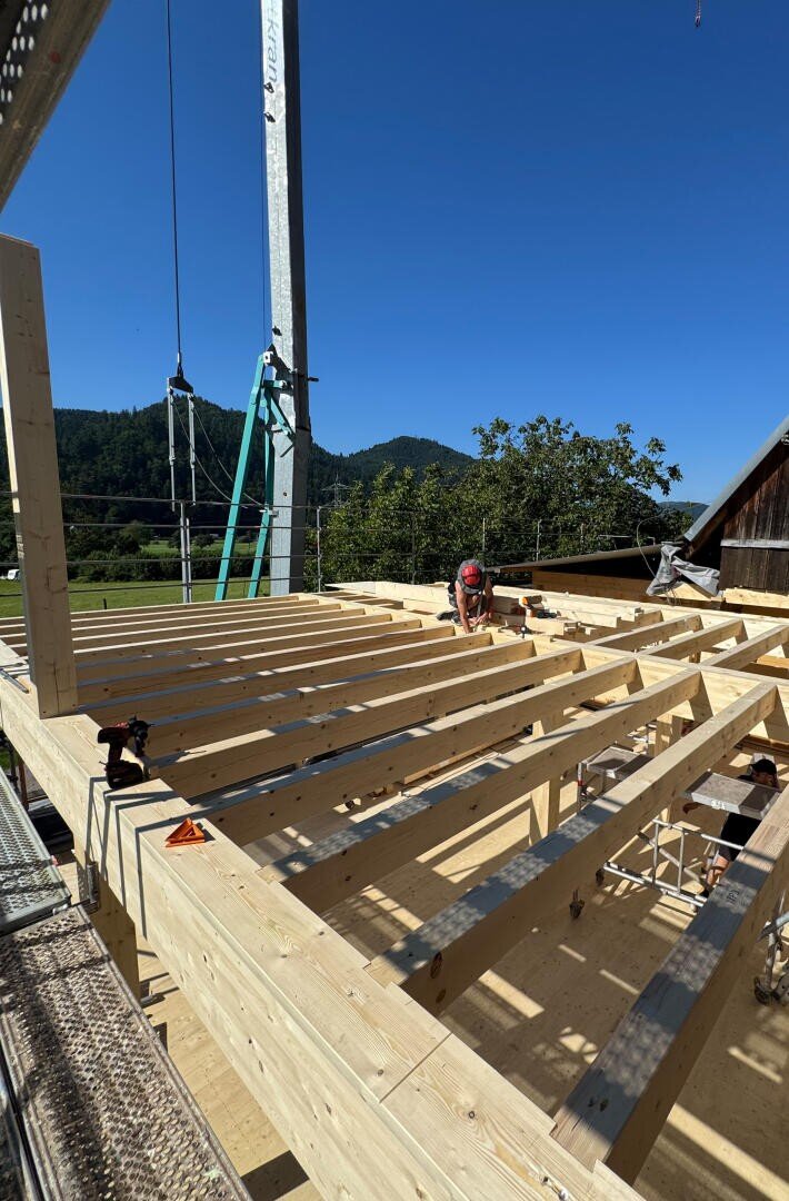 Ein Bauarbeiter, der einen Helm trägt, kniet auf einem Holzgerüst eines Gebäudes unter blauem Himmel, mit grünen Hügeln und Bäumen im Hintergrund. Bauwerkzeuge und -geräte liegen verstreut herum.