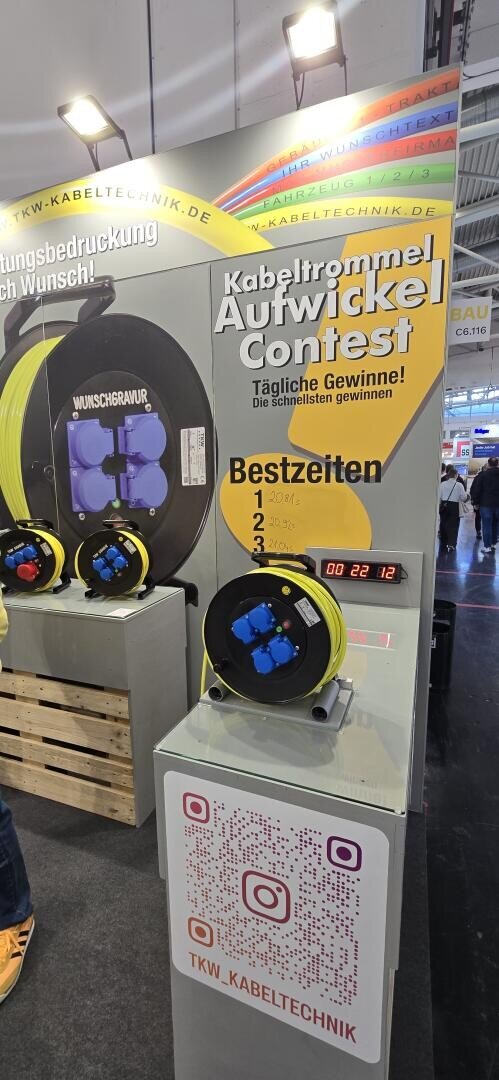 Ein Display auf einer Messe zeigt einen Kabeltrommel Aufwickel Contest mit Timer, gelber Kabeltrommel, Leaderboard und QR-Code. Der Stand wirbt für TKW-Kabeltechnik und lädt die BesucherInnen zum Mitmachen und Gewinnen ein.